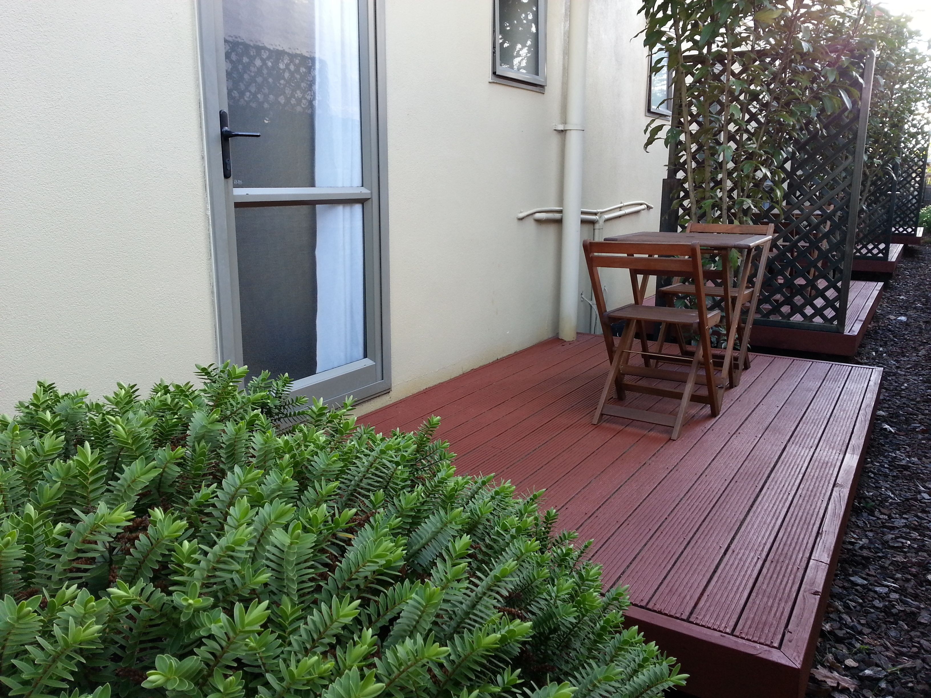A wooden deck with a table and chairs, surrounded by a hedge and a lattice fence.
