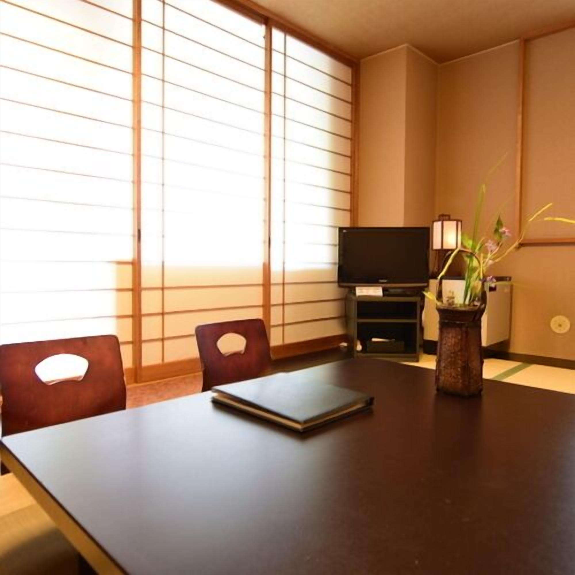A room with a dark wooden table, two chairs, a television on a stand, and a vase with flowers.