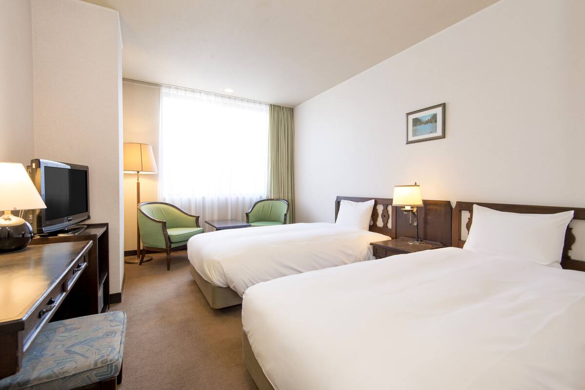 A hotel room with two beds, a television, a desk with a lamp, a chair, and a window with curtains.