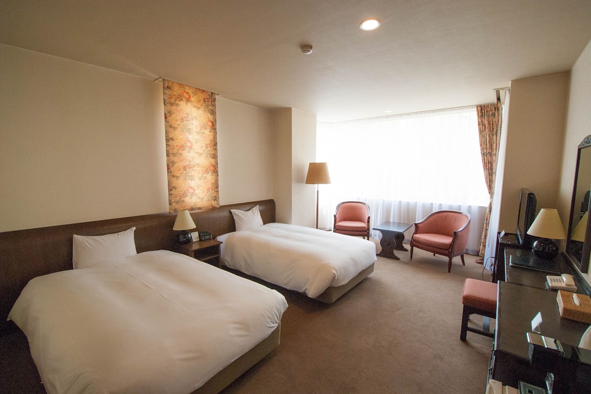 A hotel room with two beds, a desk, a chair, and a large window with curtains.