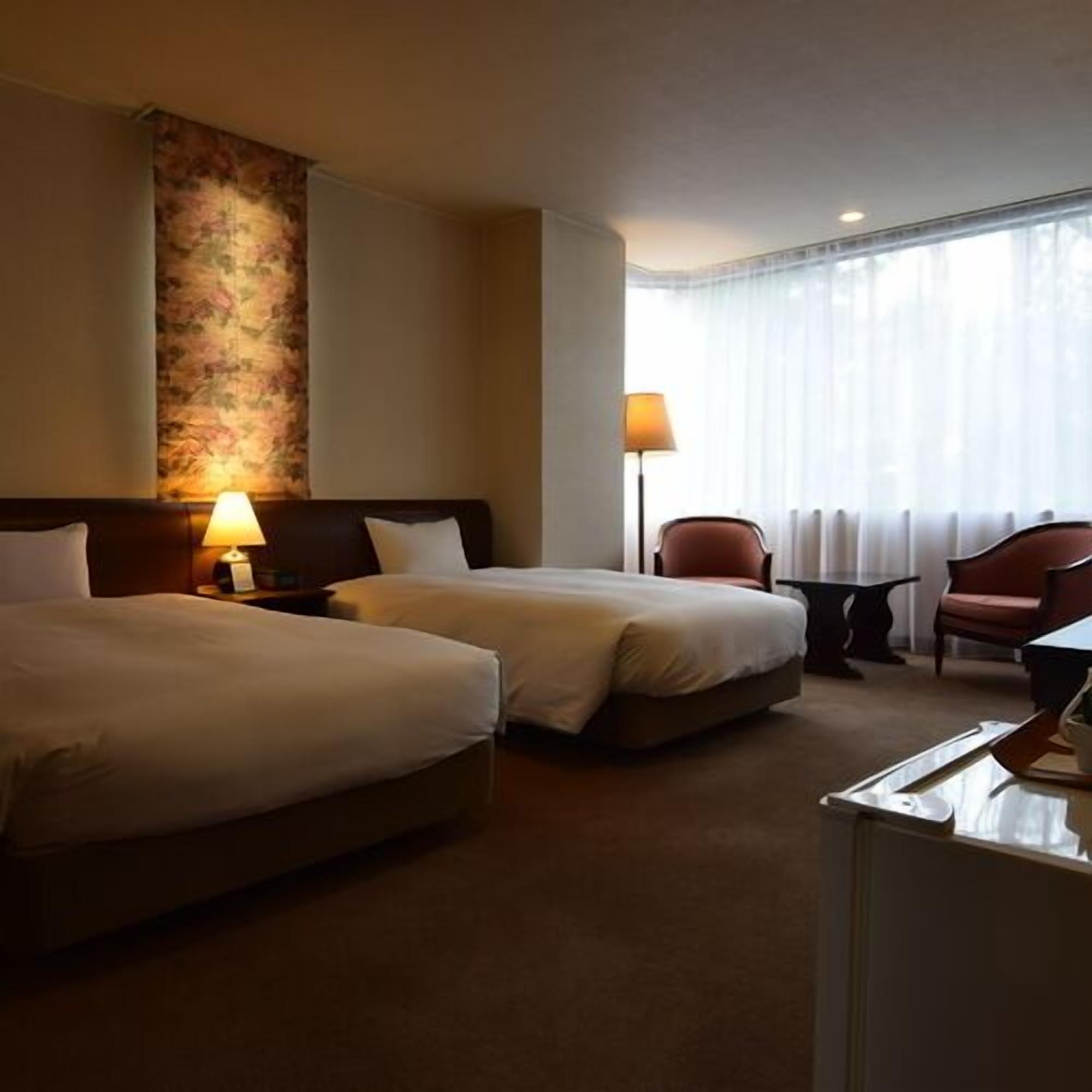 A hotel room with two beds, a desk, a chair, and a lamp.