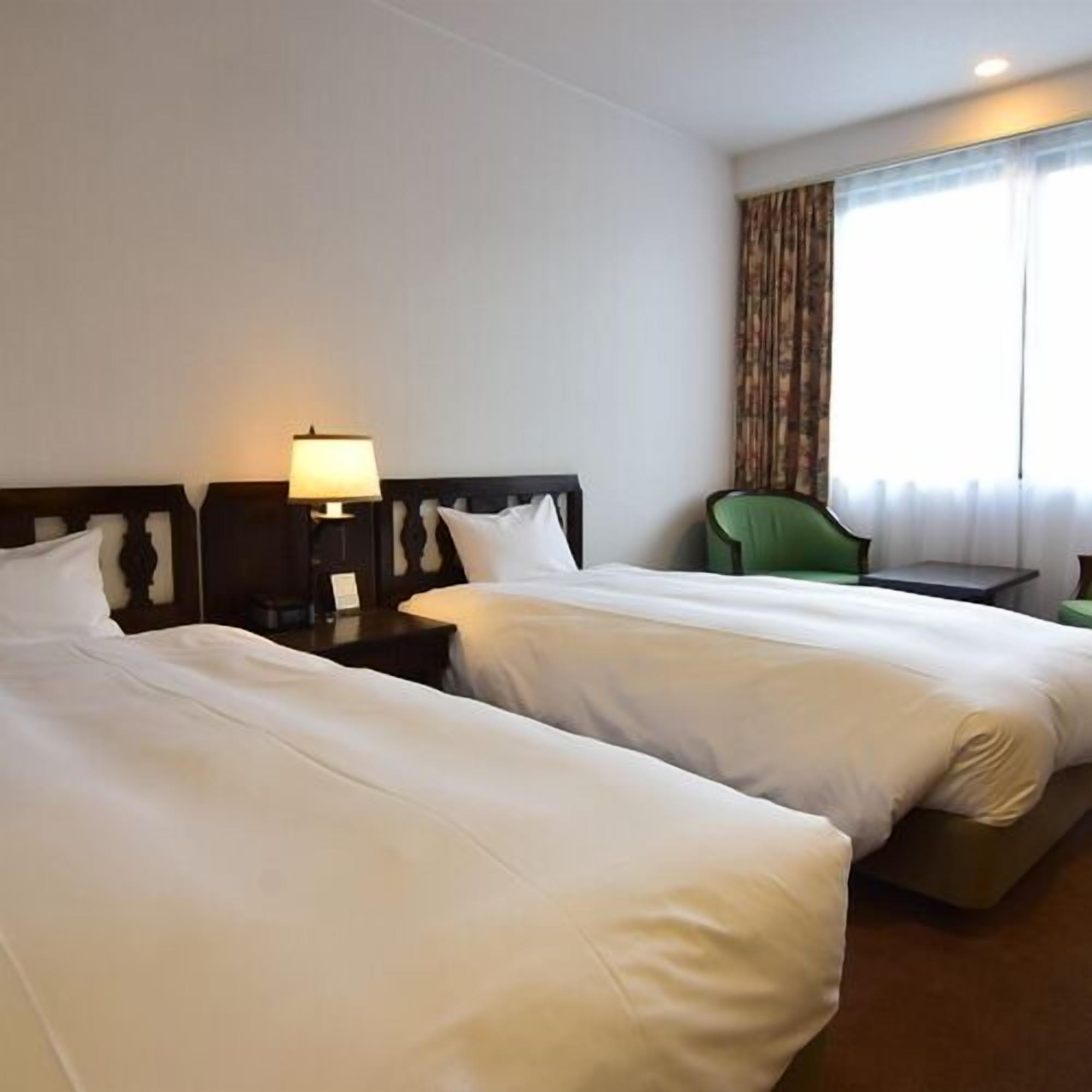 A hotel room with two beds, a nightstand with a lamp, a green chair, and a window with curtains.