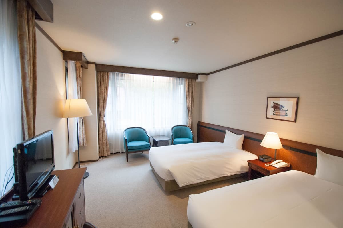 A hotel room with two beds, a desk with a TV, a chair, a bedside table with a lamp, and a window with curtains.