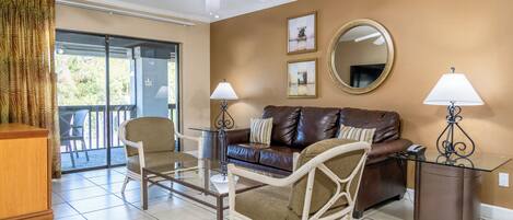 A living room with a glass coffee table, two chairs, a sofa, a ceiling fan, a mirror, and a view through a glass door.