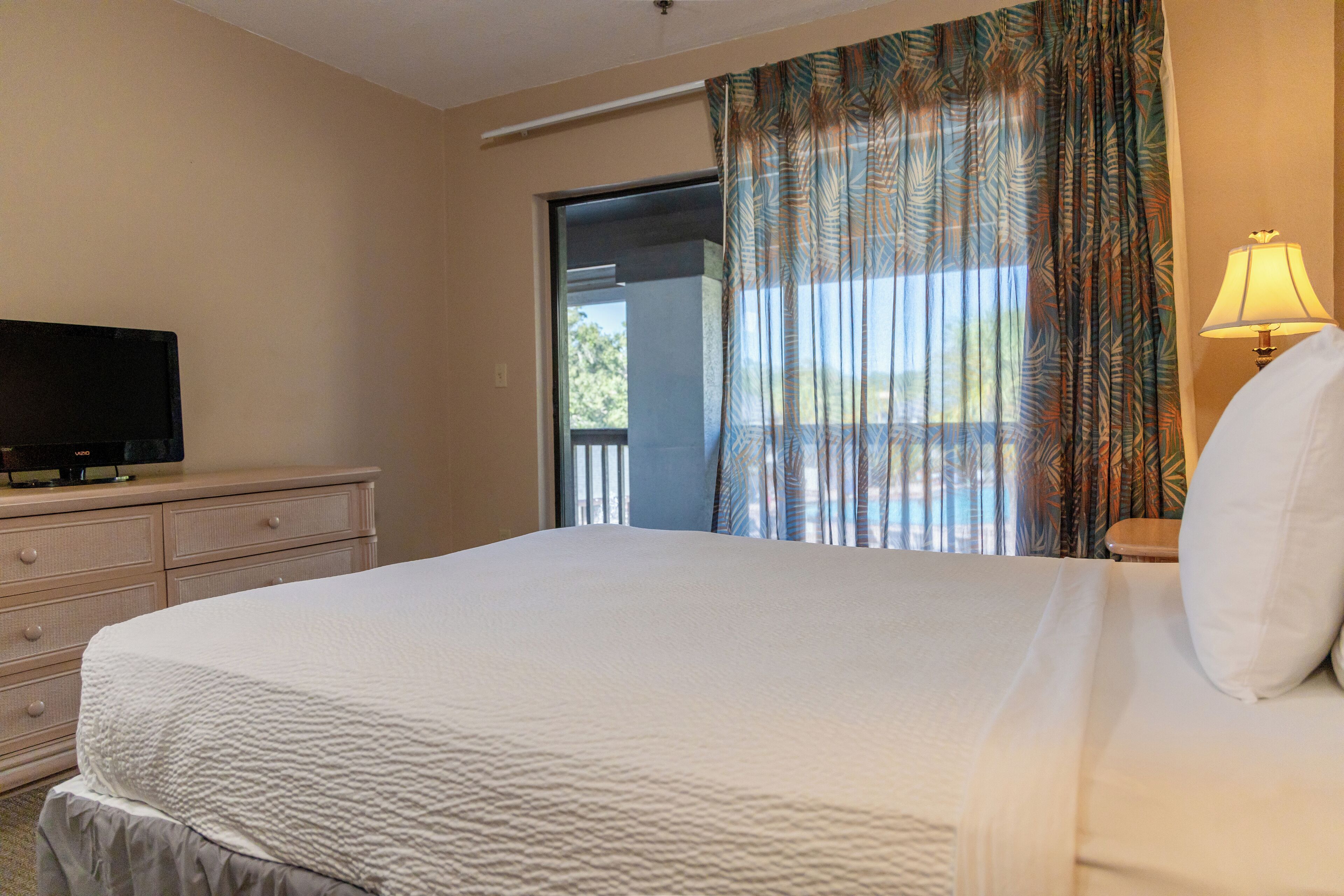 A hotel room with a bed, a television on a dresser, a lamp, and a view of the outdoors through a window with curtains.