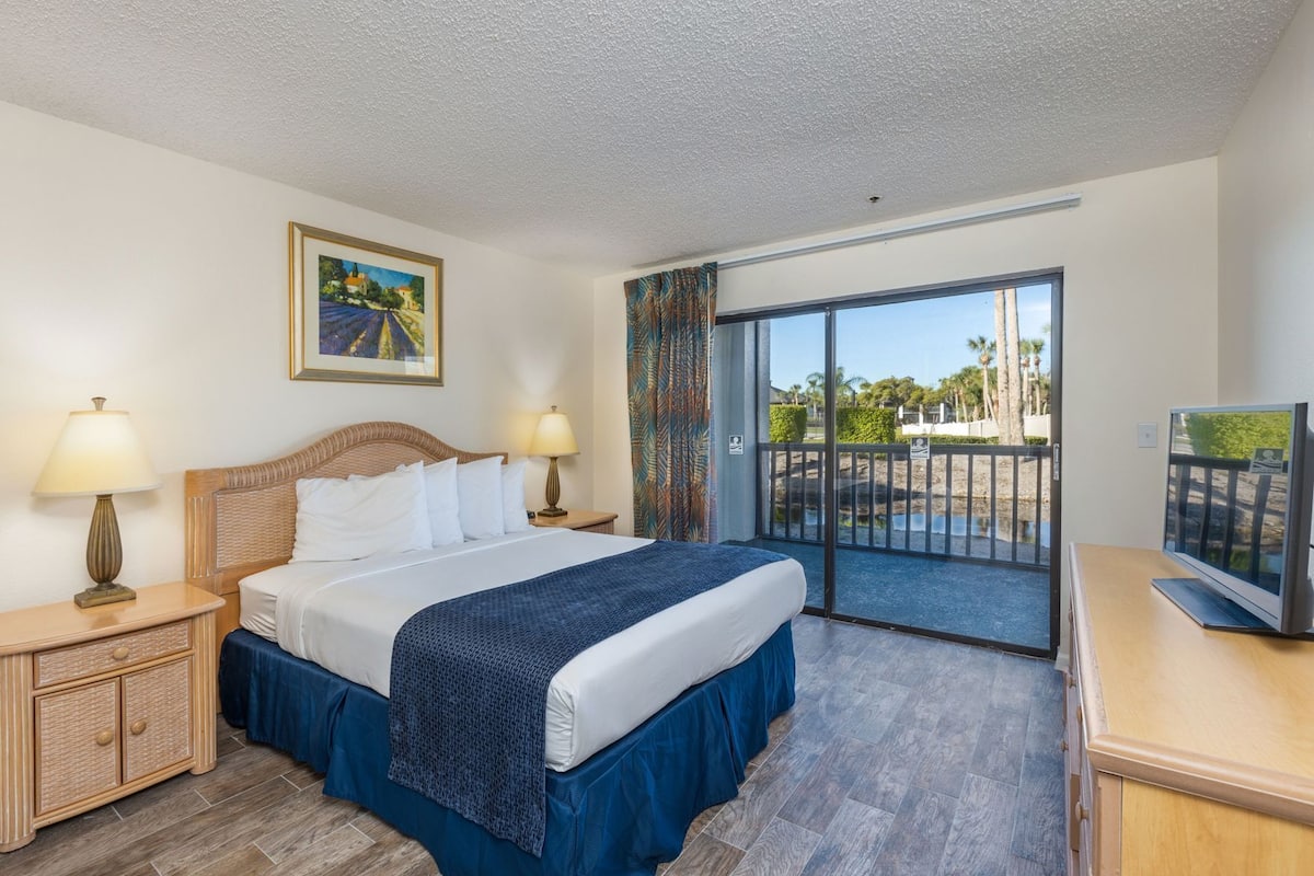A hotel room with a bed, two bedside lamps, a television, a balcony with a view of palm trees, and a painting on the wall.