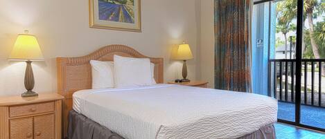 A hotel room with a large bed, two bedside tables with lamps, a painting on the wall, and a view of palm trees through the window.