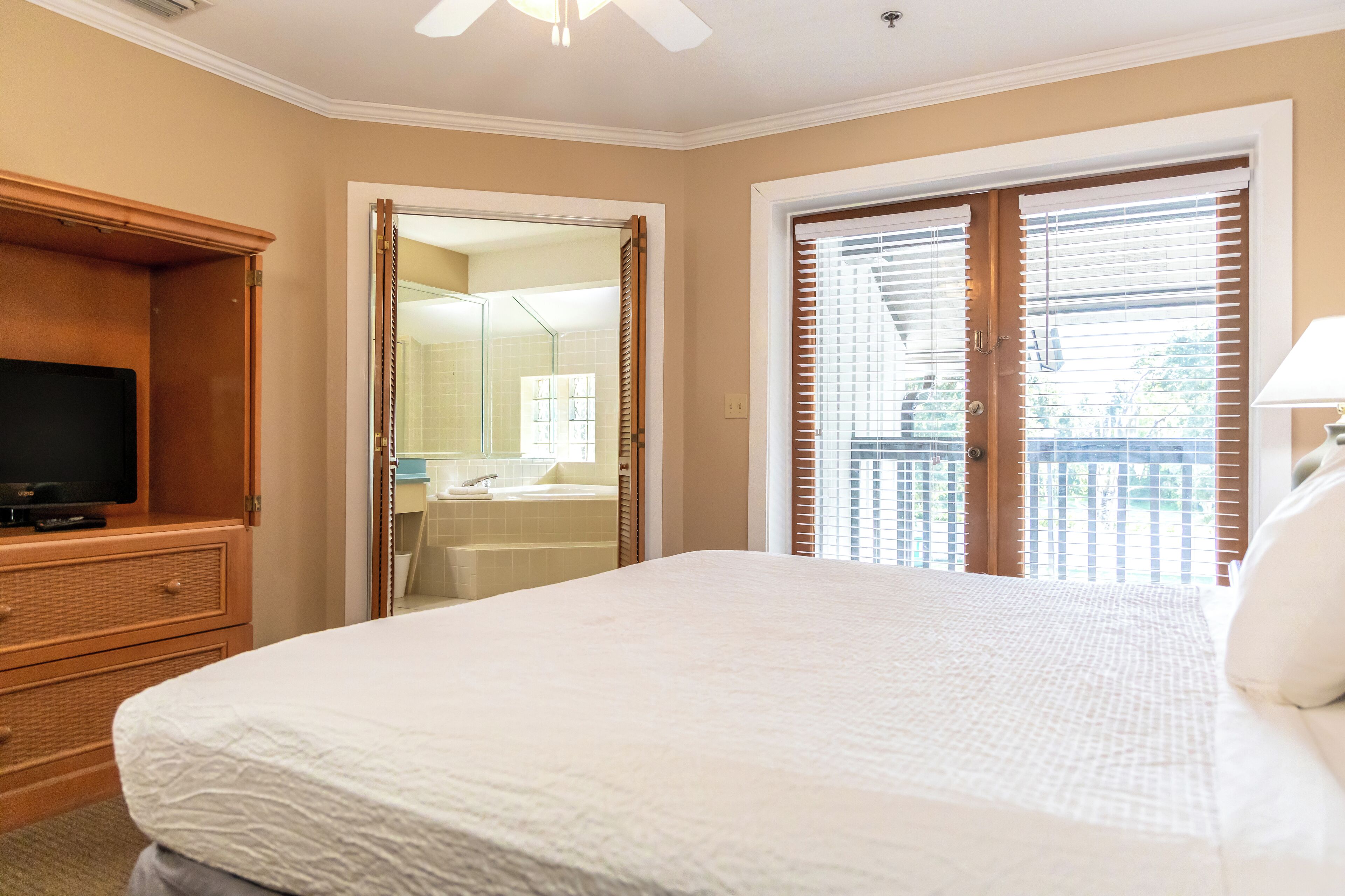 Une chambre d’hôtel avec un grand lit, un téléviseur posé sur une commode en bois, une salle de bain avec un lavabo et un miroir, et un balcon équipé de stores.