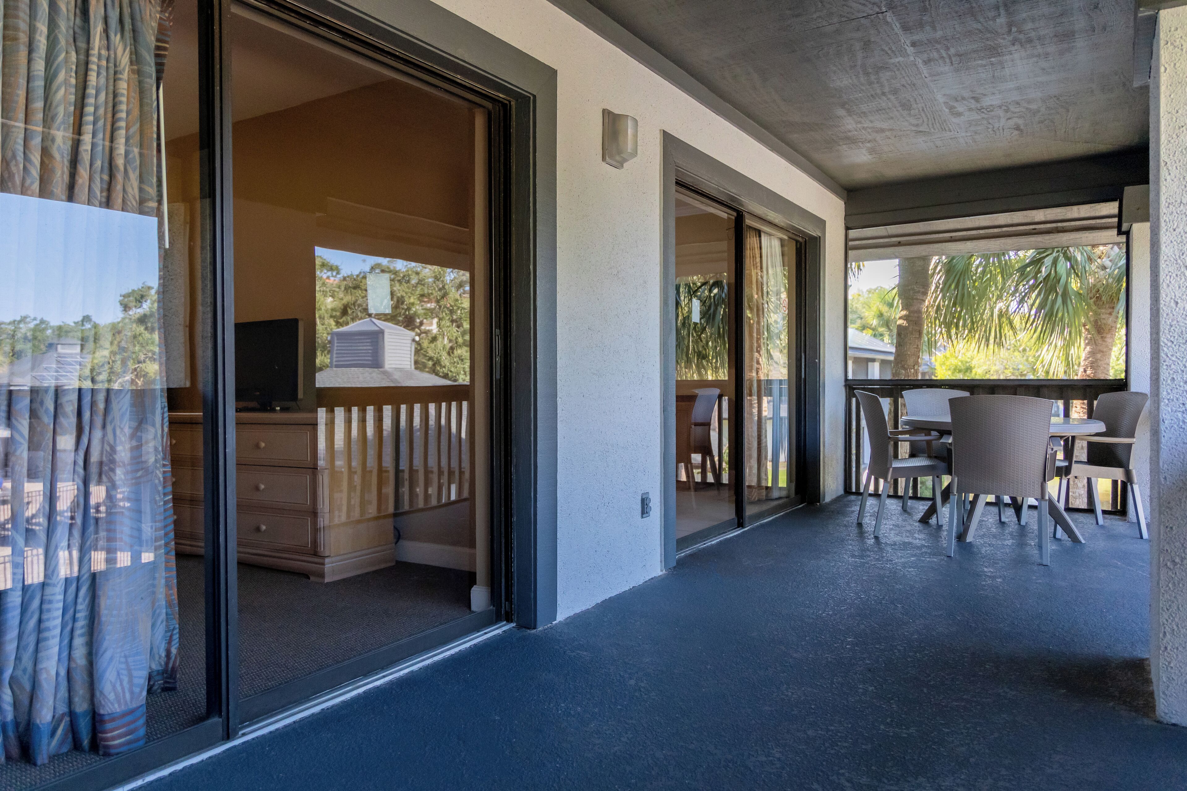 Un espacio exterior techado con una mesa y sillas de comedor, una puerta corrediza de vidrio que da a una habitación con televisor y vistas a palmeras y una casa.