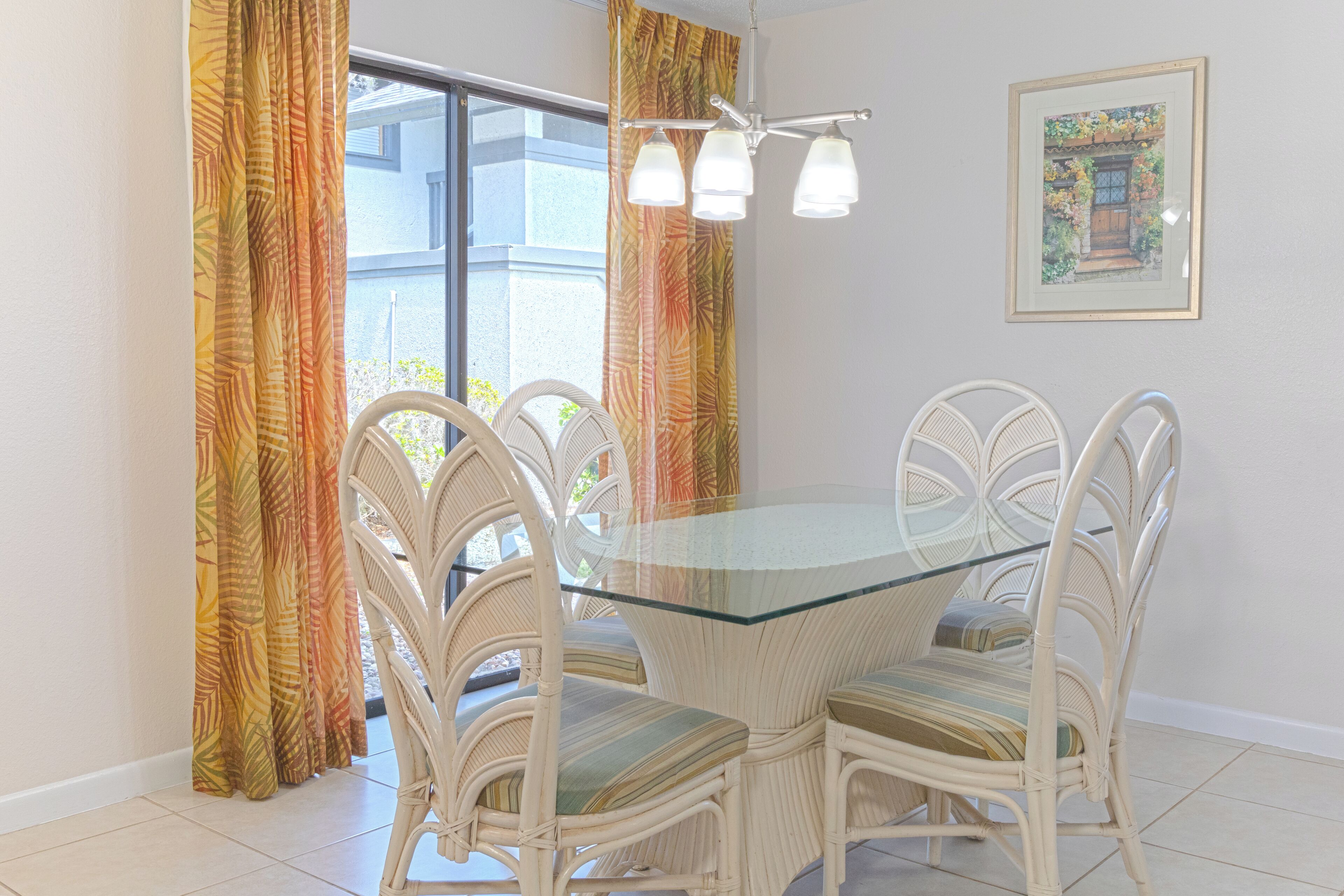 Un coin repas avec une table à plateau de verre et des chaises en osier, une grande fenêtre avec des rideaux et un tableau encadré au mur.