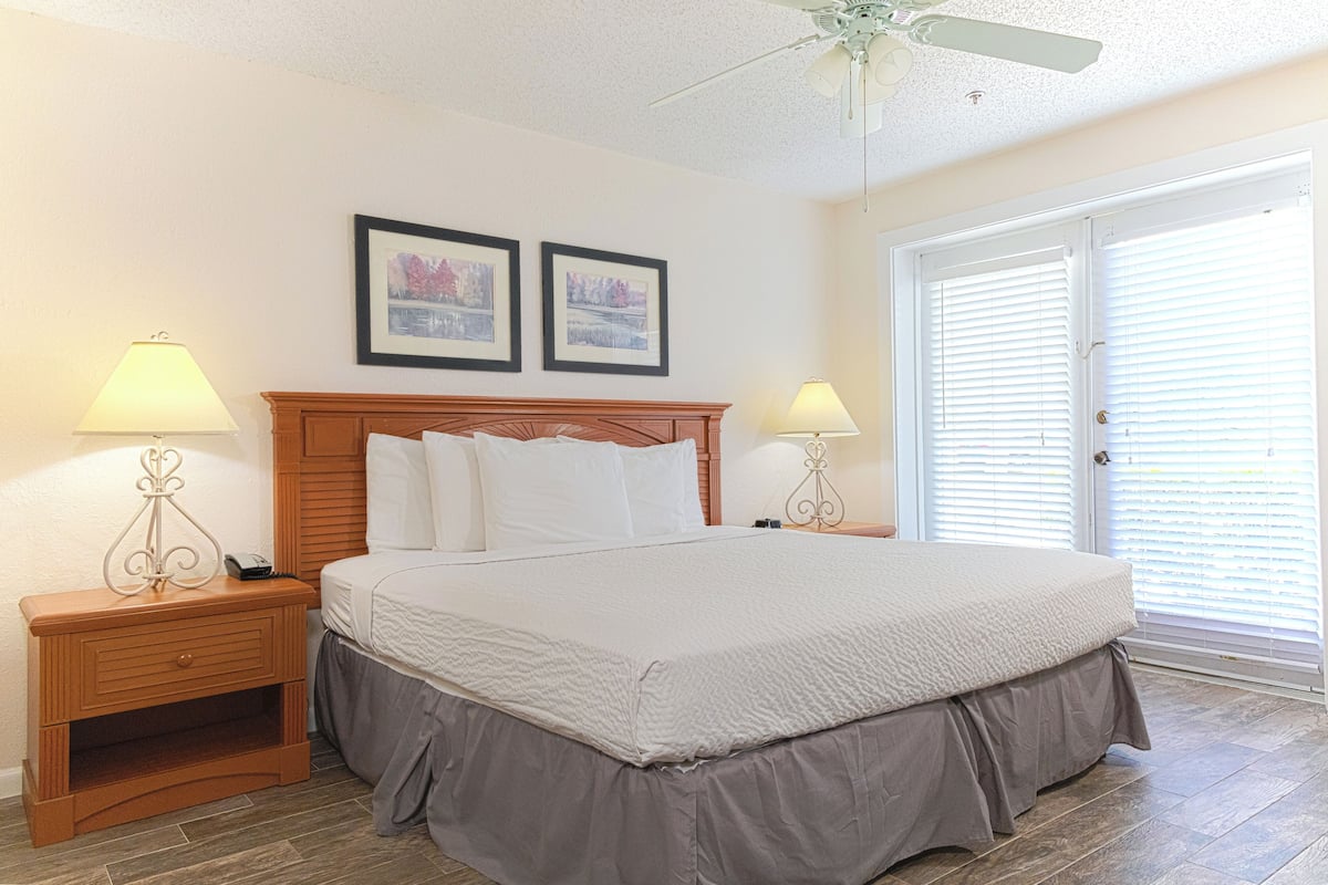 A bedroom with a large bed, two bedside tables with lamps, a ceiling fan, and two framed pictures on the wall.