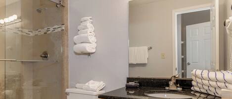 Un baño con ducha de vidrio, inodoro blanco, lavamanos con encimera de mármol y espejo con iluminación superior.