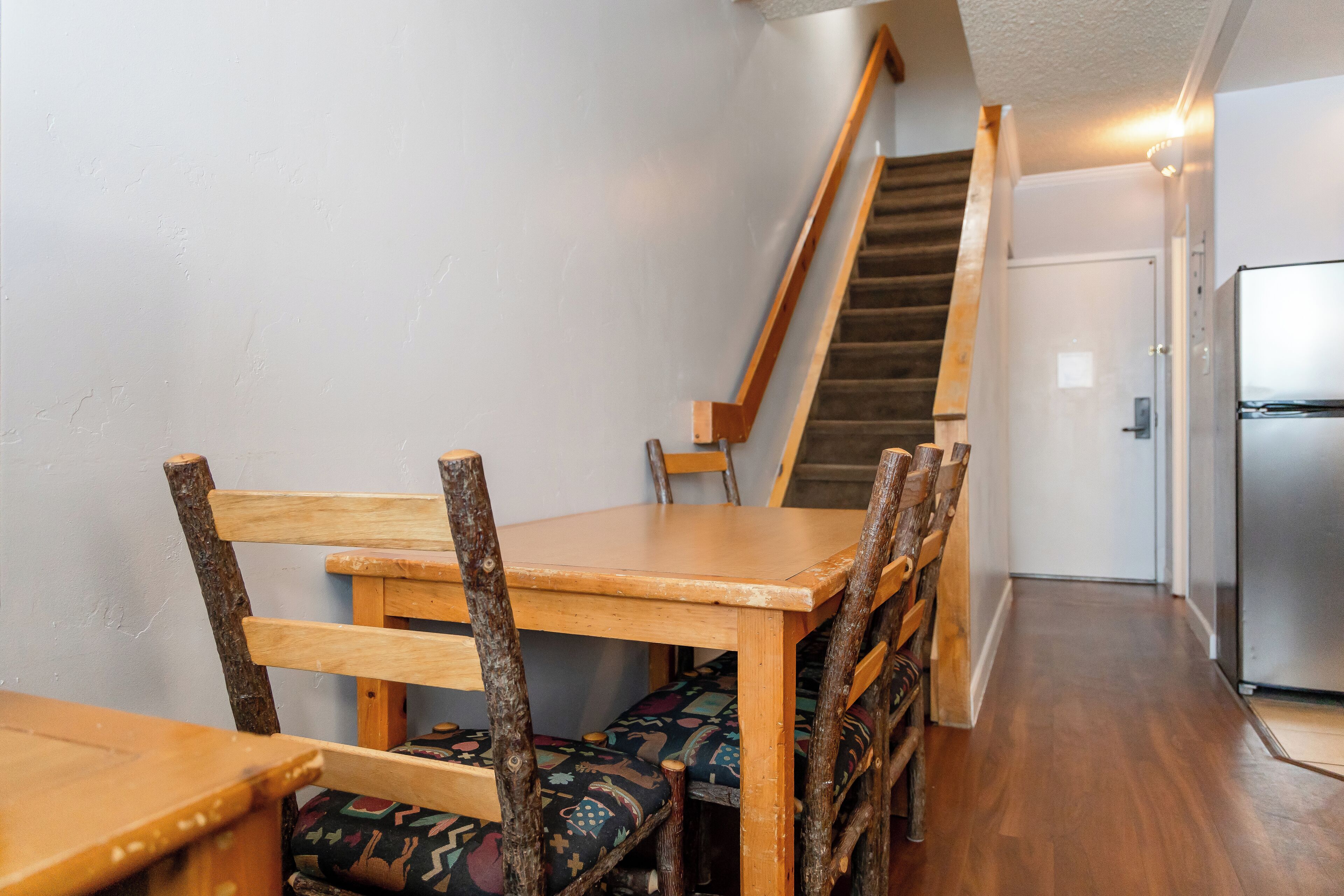 Une table à manger en bois avec des chaises, un escalier, un réfrigérateur et une porte dans un couloir.