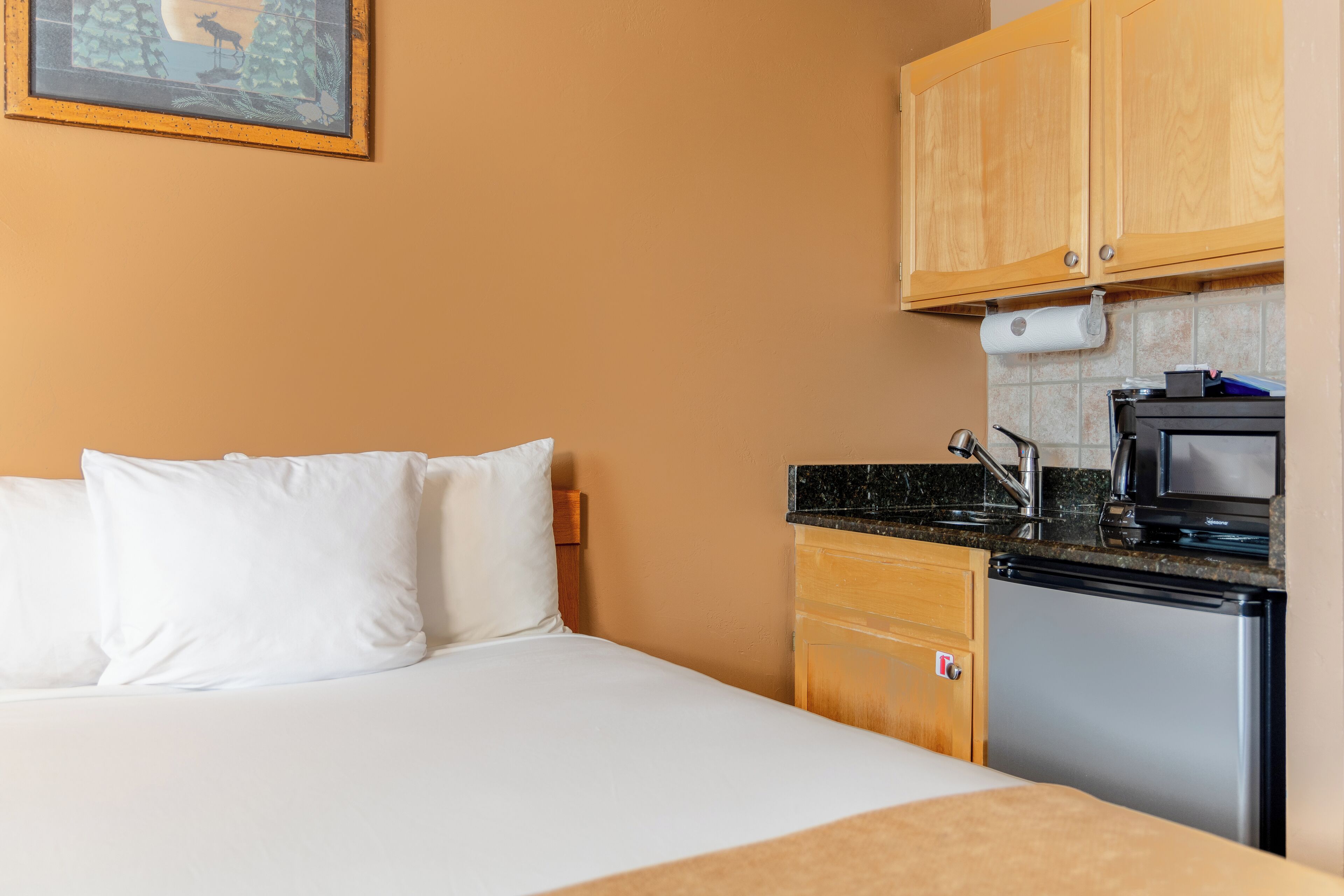 A compact hotel room with a bed, a small kitchenette, and a framed picture on the wall.
