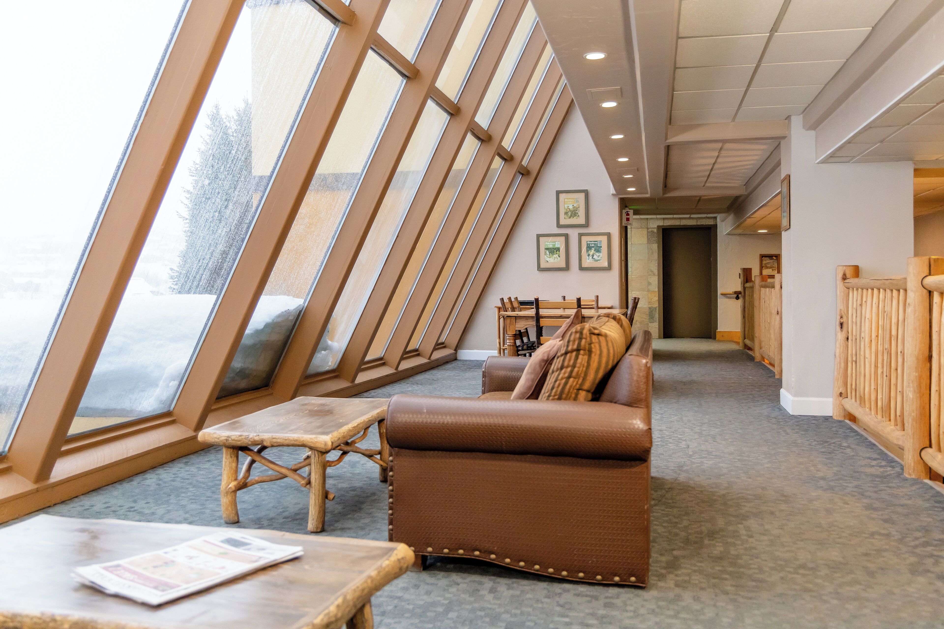 A cozy room with a sloped ceiling, a brown leather sofa, a wooden table, and a view through large windows.