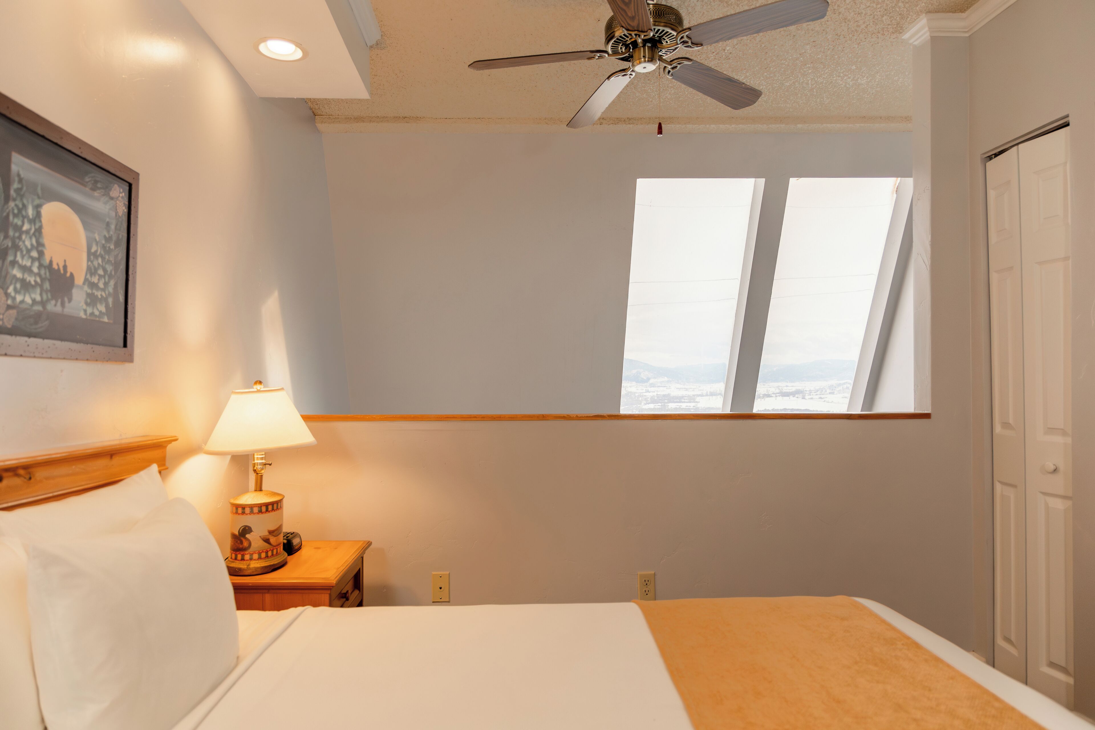 A hotel room with a bed, a nightstand with a lamp, a ceiling fan, and a window with a view.