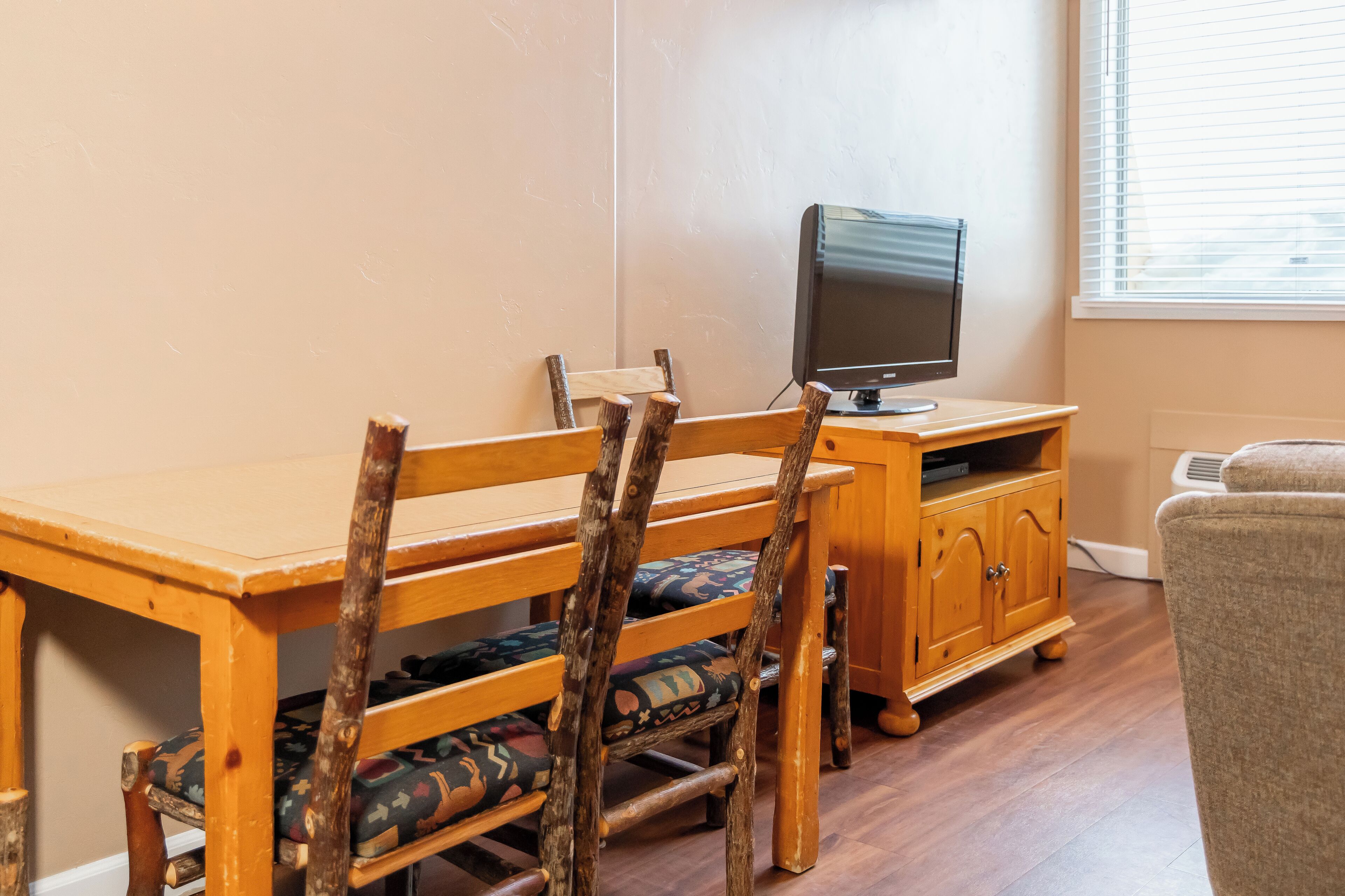 A wooden dining table with chairs, a television on a wooden stand, and a window with blinds.
