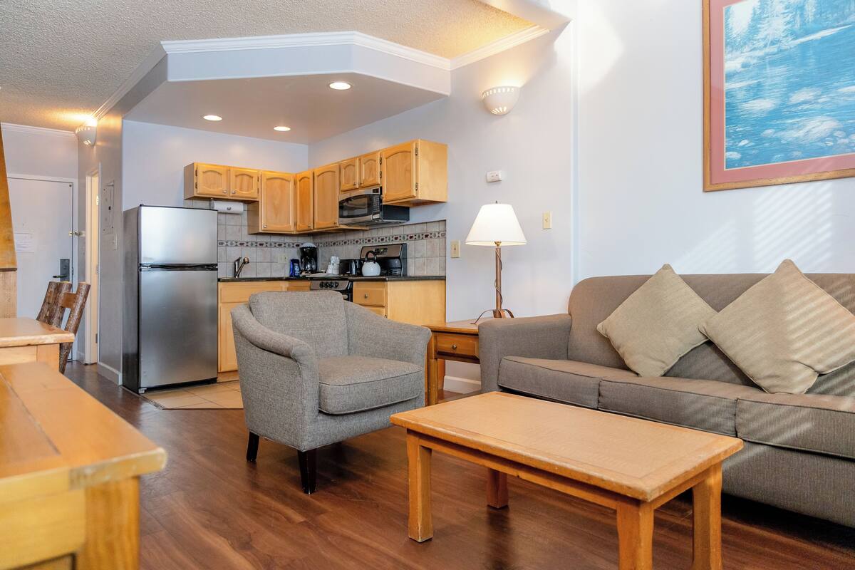 A compact living space with a kitchenette, a gray sofa, a wooden coffee table, and a gray armchair.