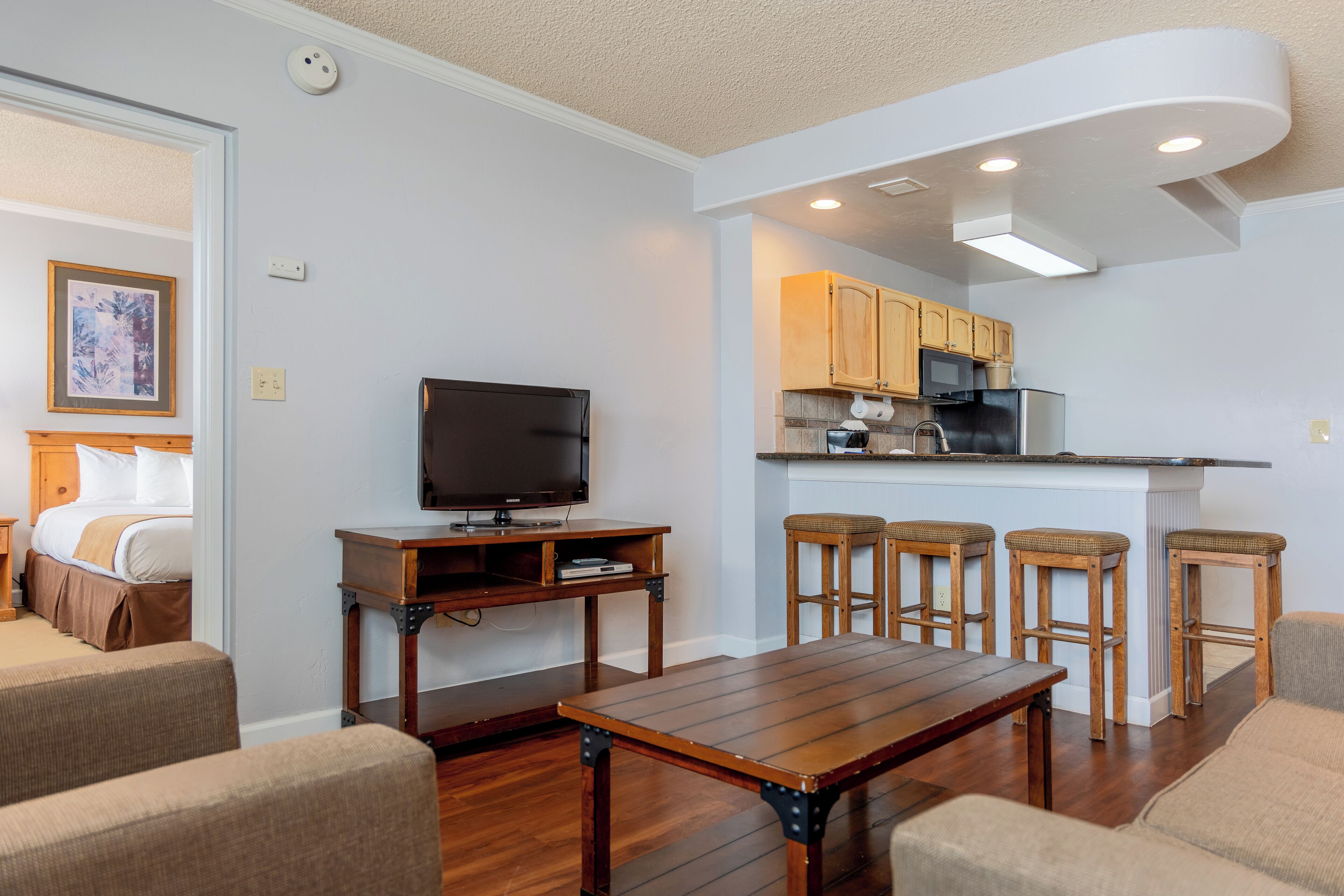 A modern living room with a wooden coffee table, a TV, and comfortable seating. Adjacent is a kitchen with wooden cabinets and a bar area.