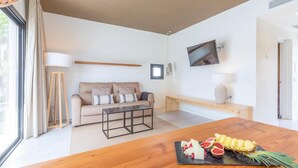 A modern living room with a sofa, a coffee table, a wooden dining table with fruit, a TV, and a large window with curtains. - Marsenses Paradise Club Hotel (Ciutadella de Menorca)