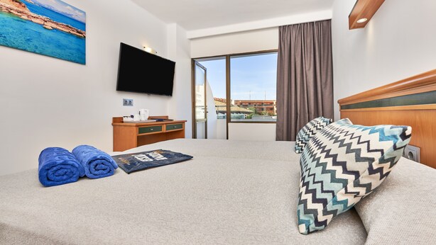 Habitación de hotel con cama, escritorio, televisión de pantalla plana, un cuadro y ventana con vista a edificios.