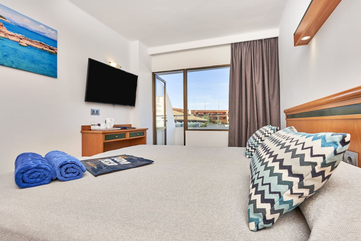 A hotel room with a bed, a desk, a flat-screen TV, a painting, and a window with a view of buildings.