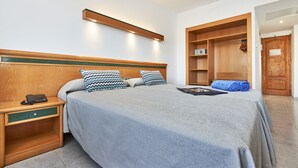 Una cama bien hecha con cubrecama gris y dos almohadas estampadas, una mesita de noche de madera con un teléfono y un armario de madera con una toalla encima.