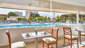 Zona de comedor junto a la piscina con mesas y sillas, con vistas a una piscina y al paisaje urbano.