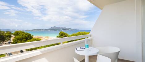 Un balcón con una mesa y sillas blancas, con vista a una playa y montañas.