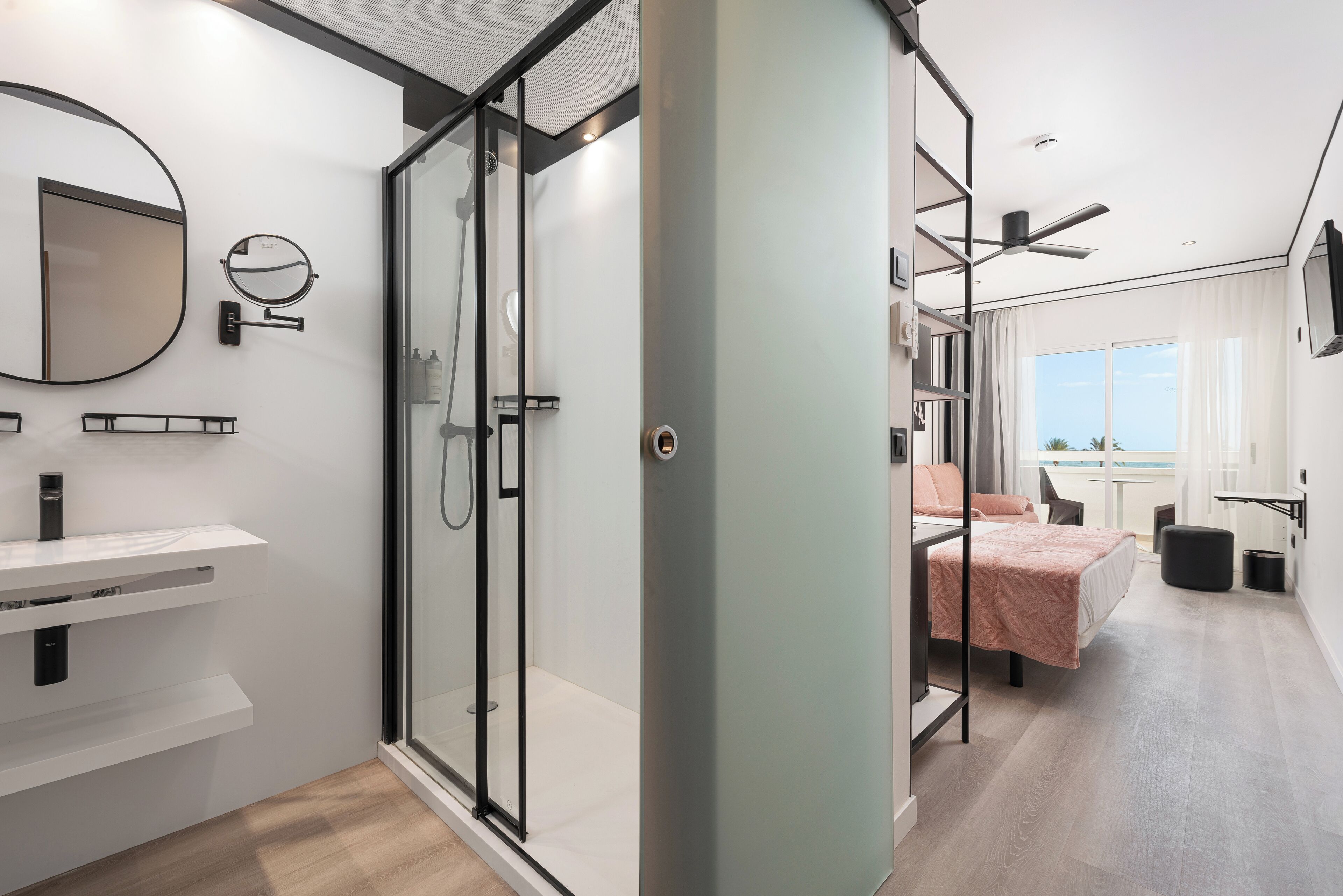Una habitación de hotel moderna con cabina de ducha de vidrio, cama con colcha rosa, una mesita y vistas al mar a través de amplias ventanas.