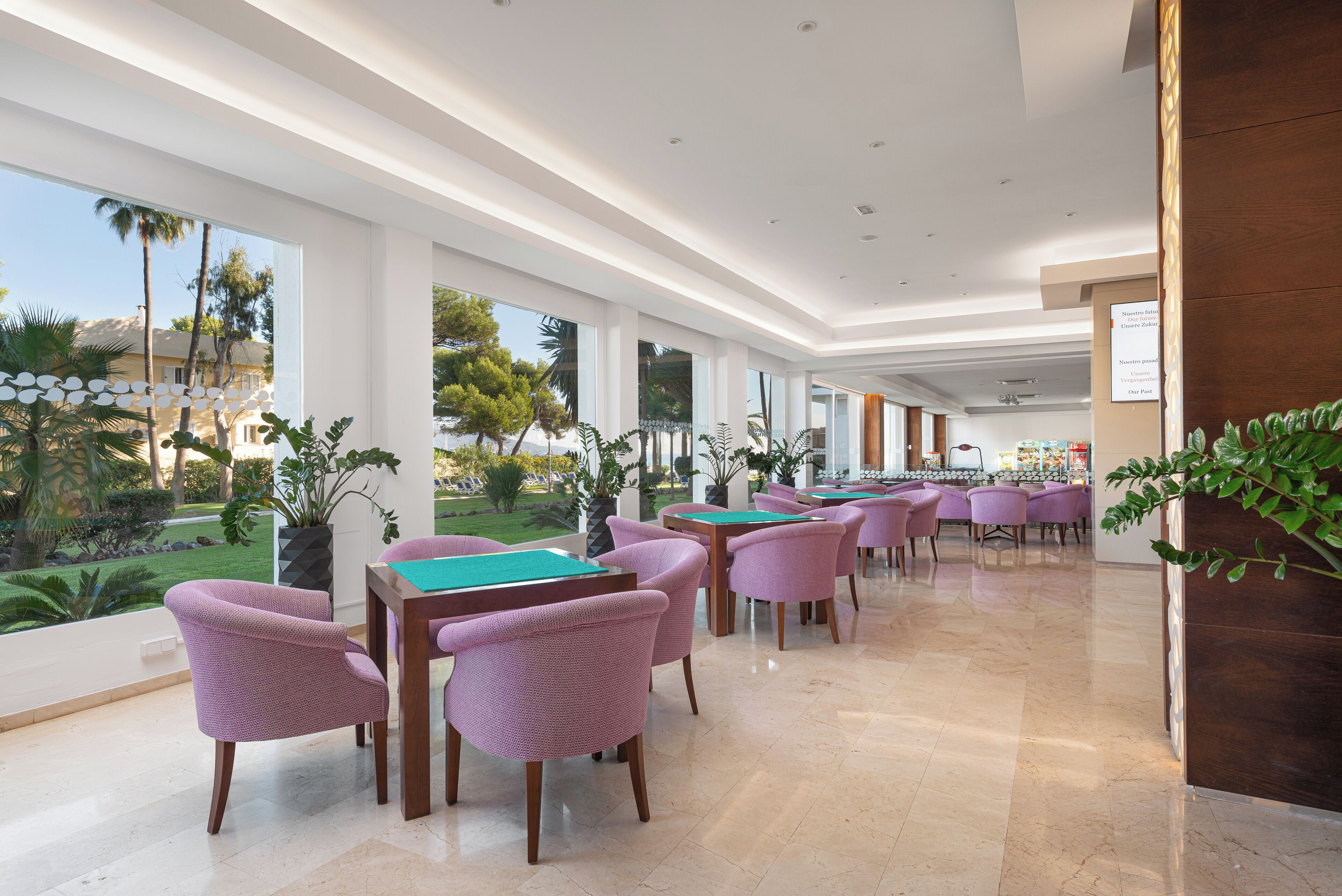 A spacious indoor area with multiple pool tables and purple chairs, large windows offering a view of a landscaped garden.