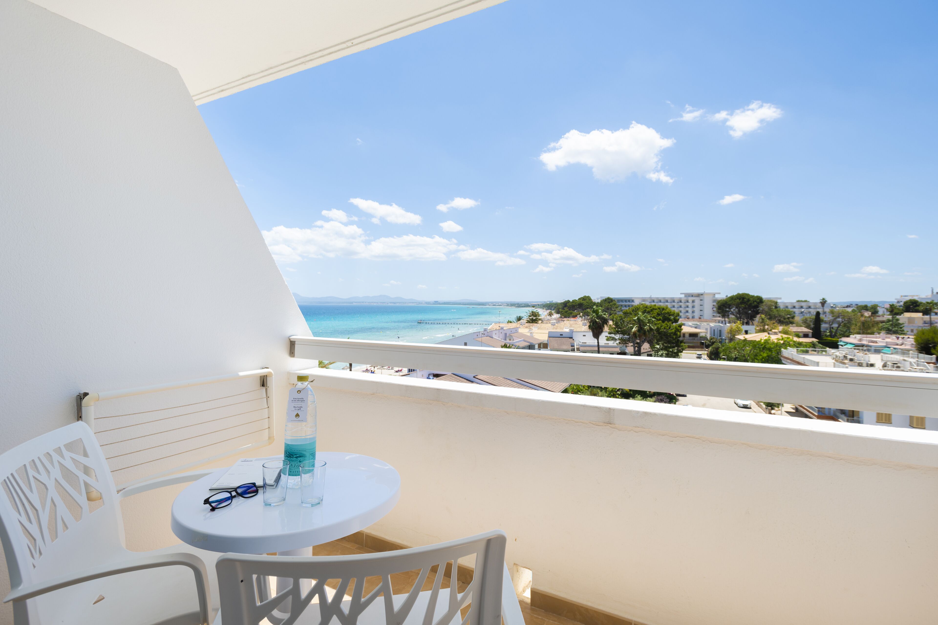 Un balcón con una mesa y sillas blancas, con vistas a la playa y al paisaje urbano.