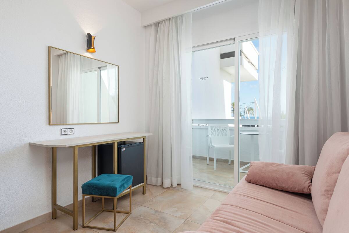 A modern hotel room with a large mirror, a small desk with a computer, a comfortable sofa, and a sliding glass door leading to a balcony.