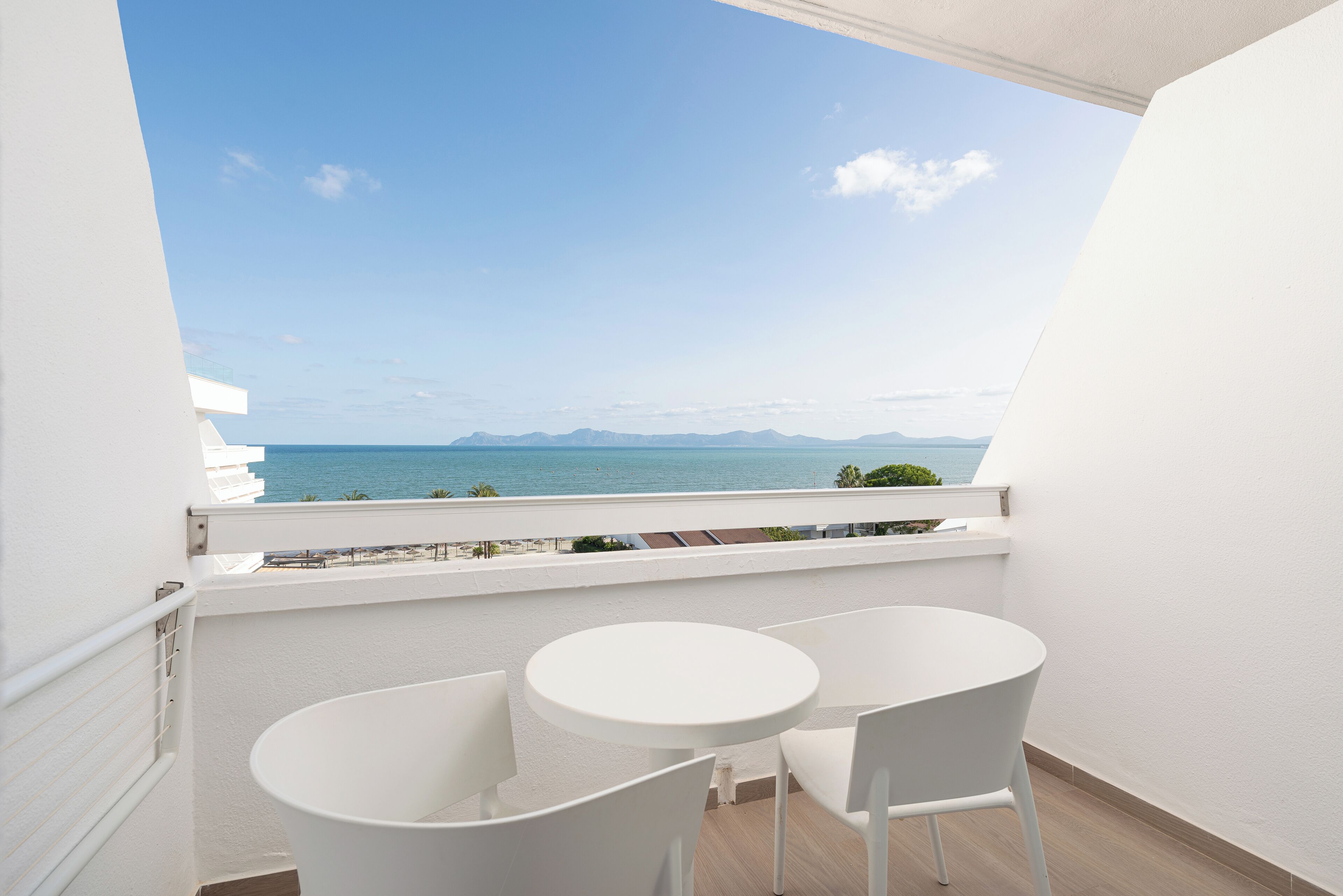 A balcony with a white table and chairs, overlooking a coastal view with mountains in the distance.