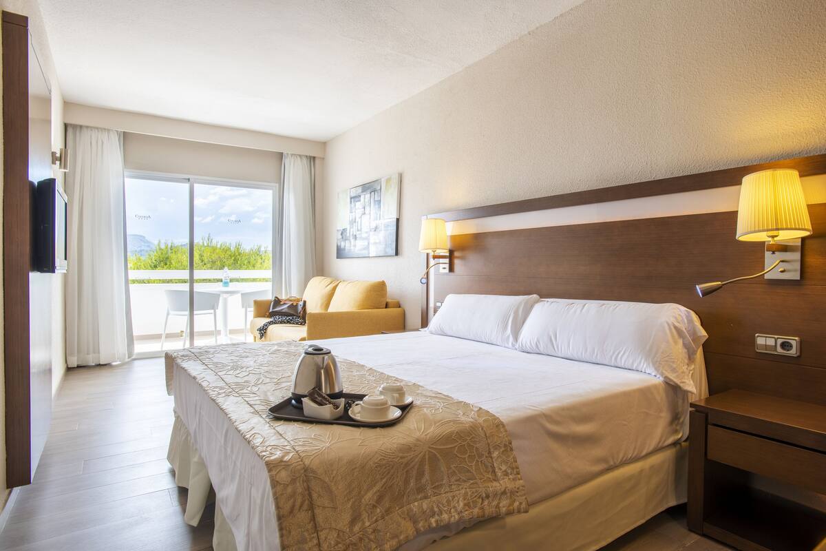 A hotel room with a bed, bedside lamps, a TV, a balcony with a table and chairs, and a coffee maker on the bed.