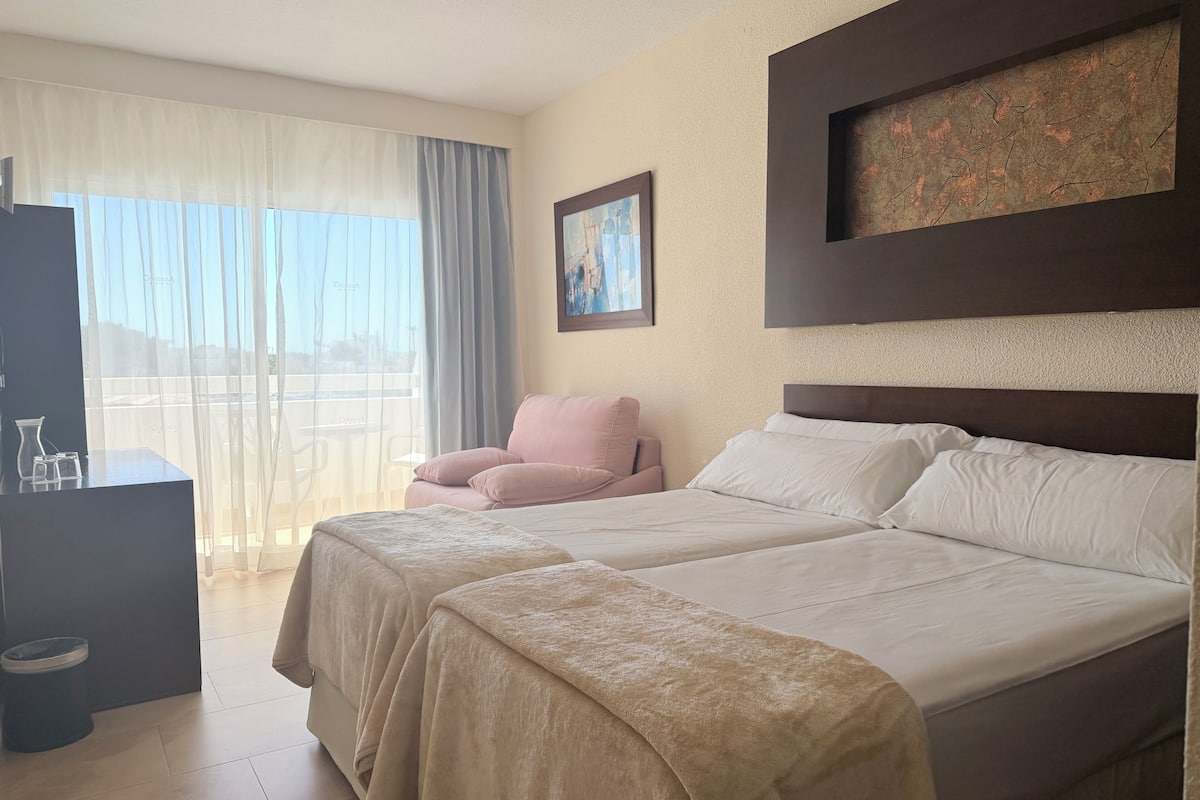 Een hotelkamer met twee bedden, een stoel, een bureau met een karaf en glazen, een schilderij en uitzicht door het raam.