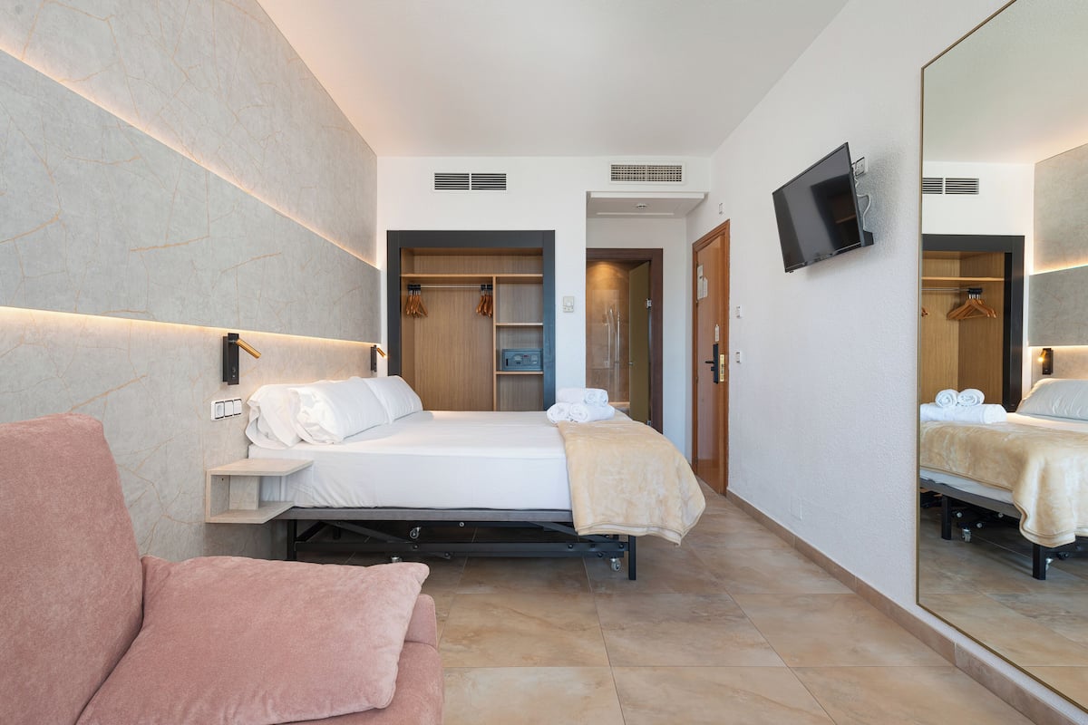 A modern hotel room with a bed, a sofa, a TV, and a large mirror.