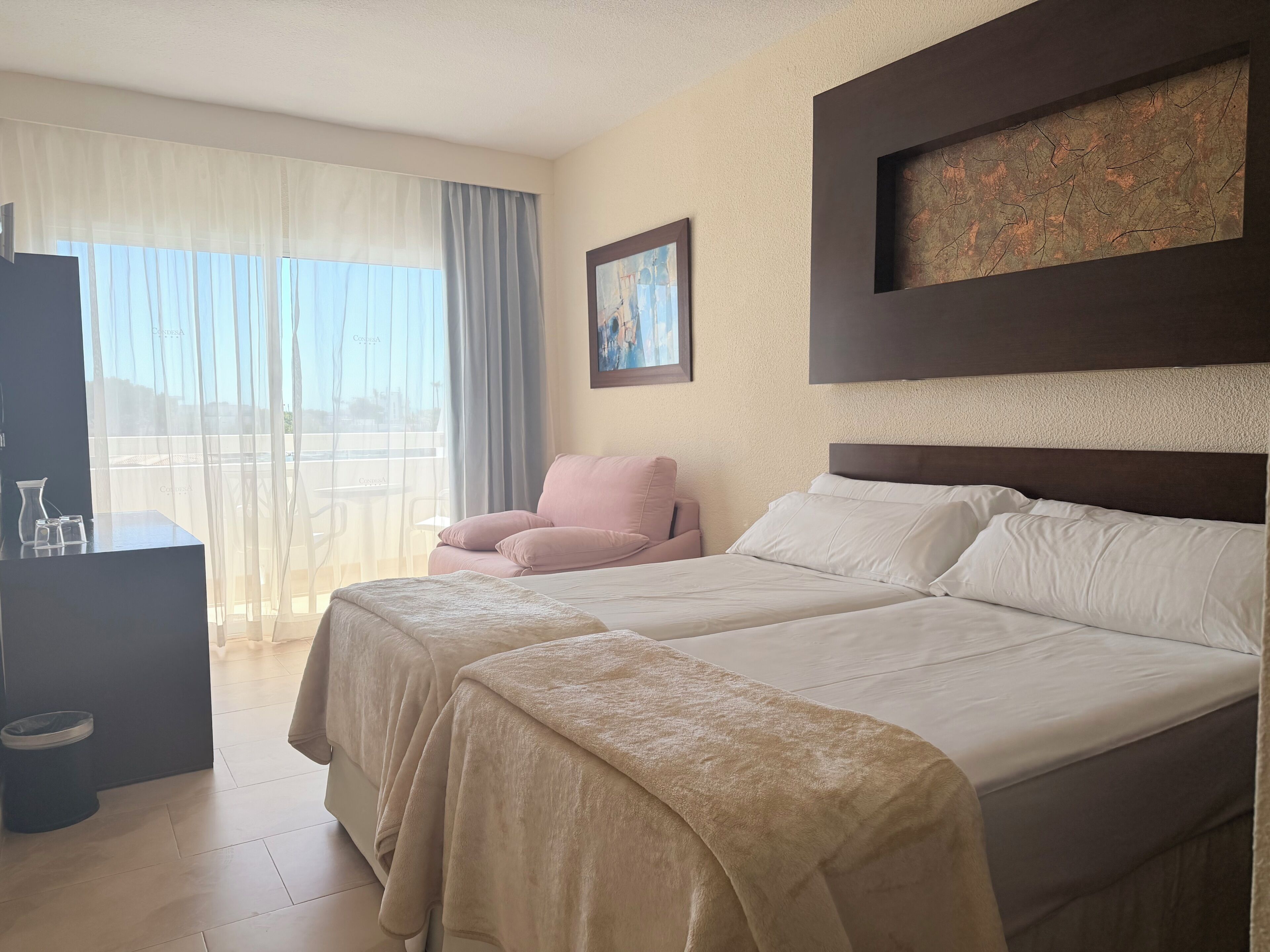 A hotel room with two beds, a chair, a desk with a pitcher and glasses, a painting, and a view through the window.