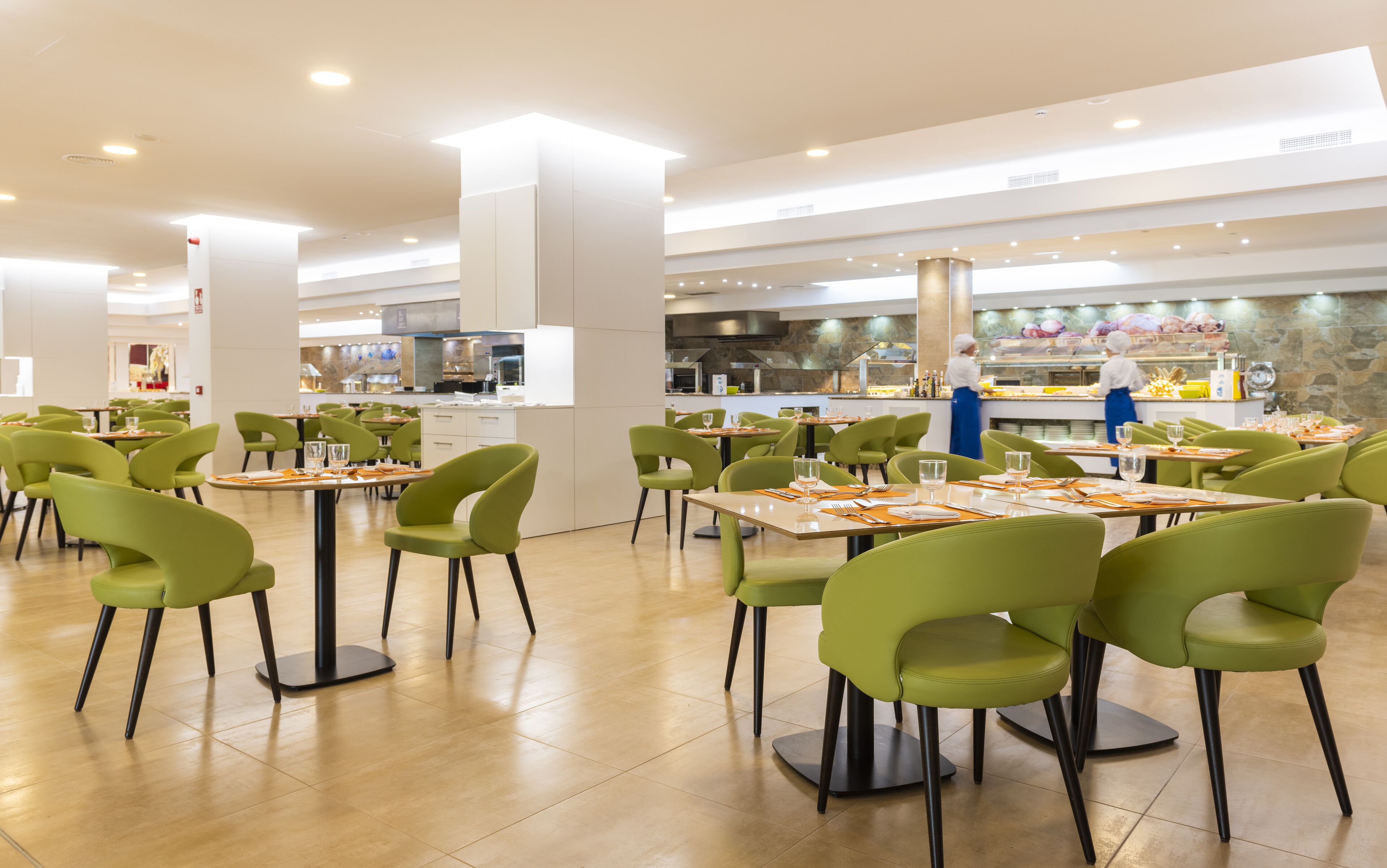 Un comedor amplio con sillas y mesas verdes, una zona de cocina central y un ambiente luminoso y bien iluminado.