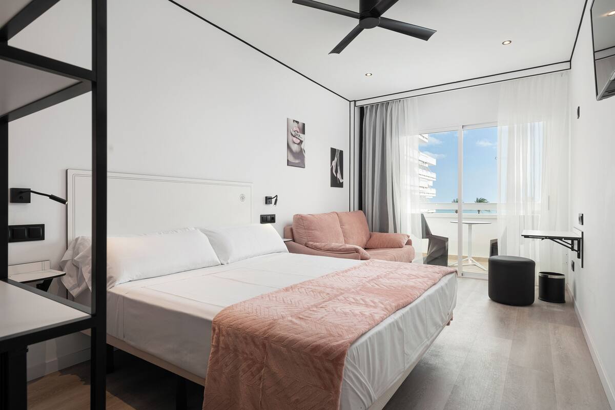 A modern hotel room with a bed, a desk, a chair, a balcony with a view, and a ceiling fan.