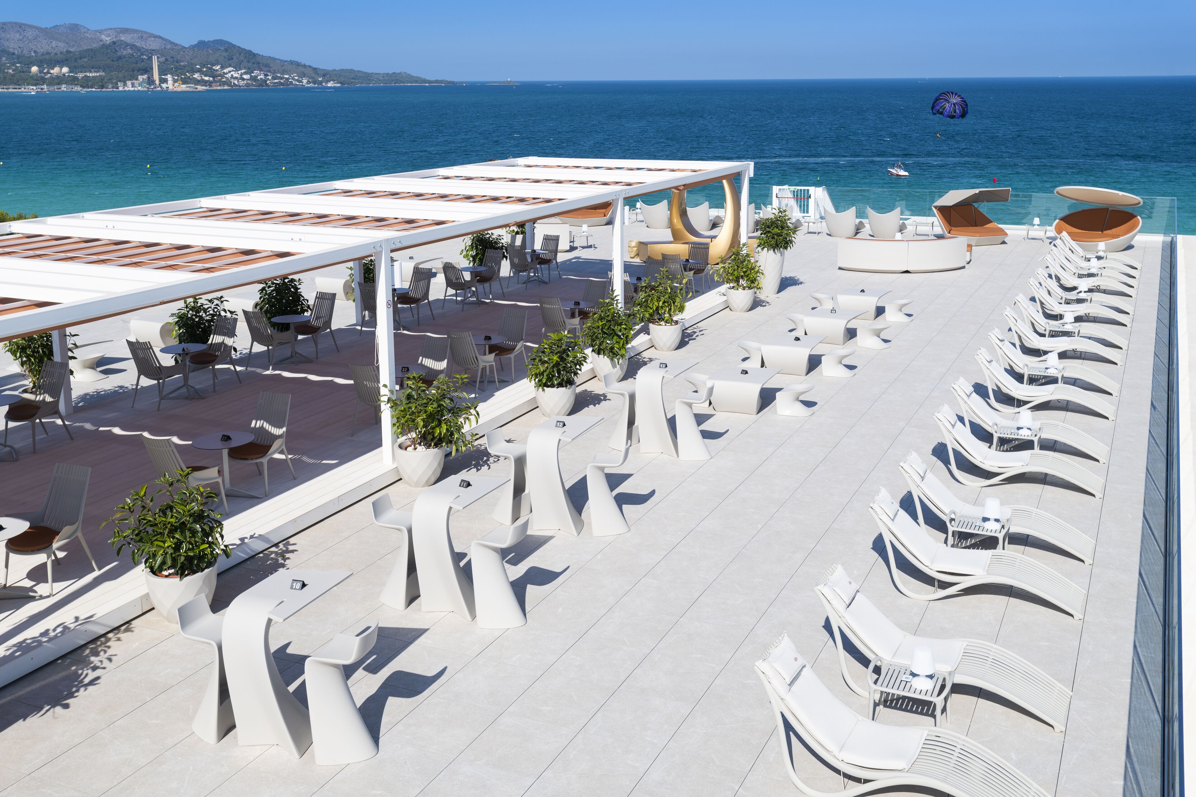 A rooftop terrace with white furniture, potted plants, and a view of the sea.