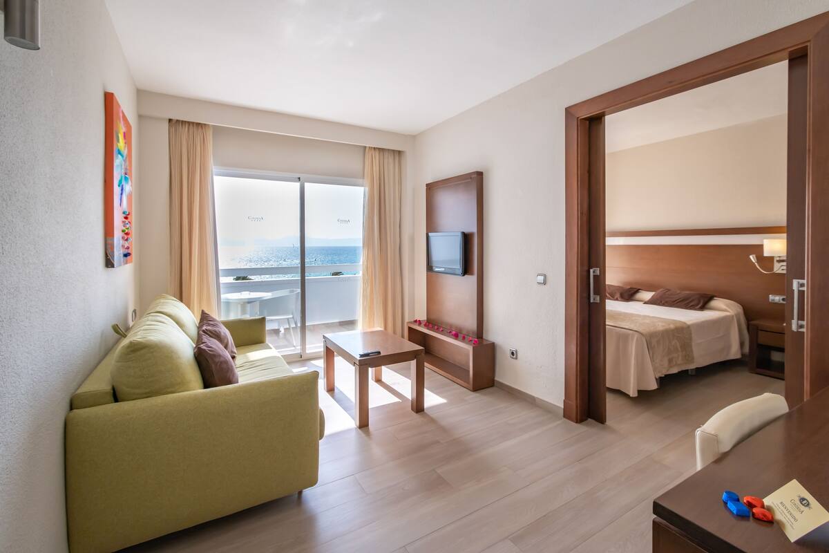 A hotel room with a sofa, a TV mounted on the wall, a small table, and a bed with a wooden headboard.