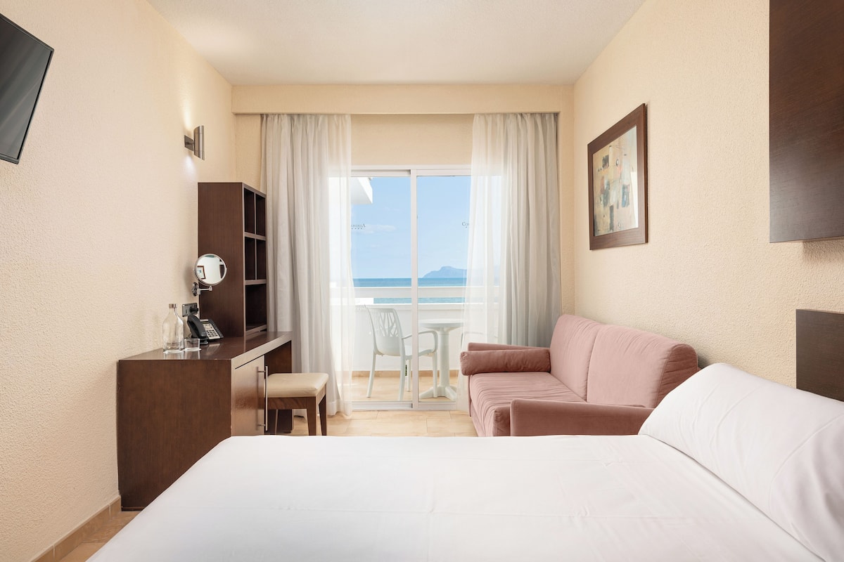A hotel room with a bed, a desk with a telephone, a chair, a sofa, a TV, and a balcony with a view of the sea.