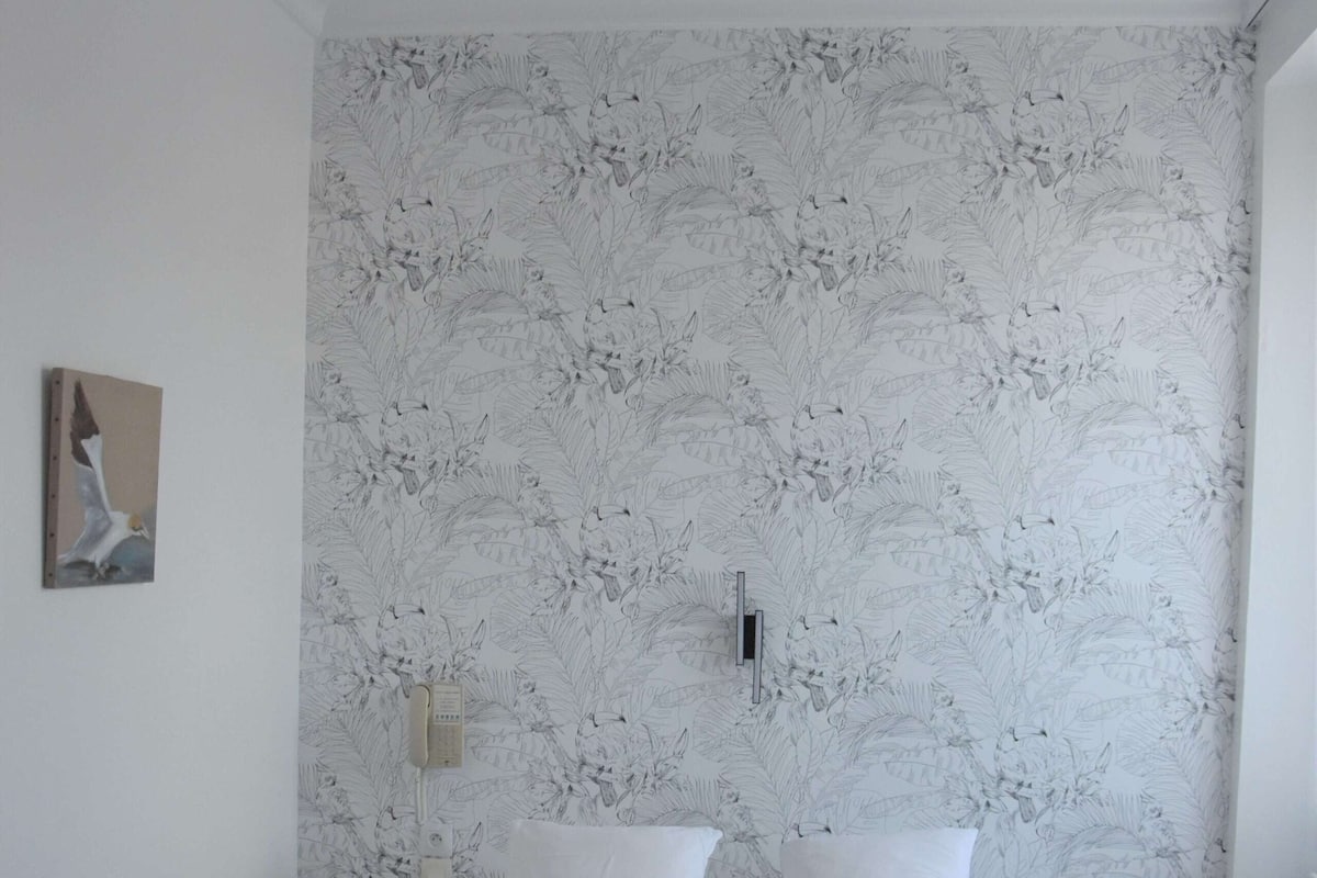 Ein ordentlich bezogenes Bett mit weißen Leinentüchern und Handtüchern, ein Deckenventilator, blumiger Wandteppich, ein Nachttisch mit Lampe und ein kleiner Bilderrahmen an der Wand.