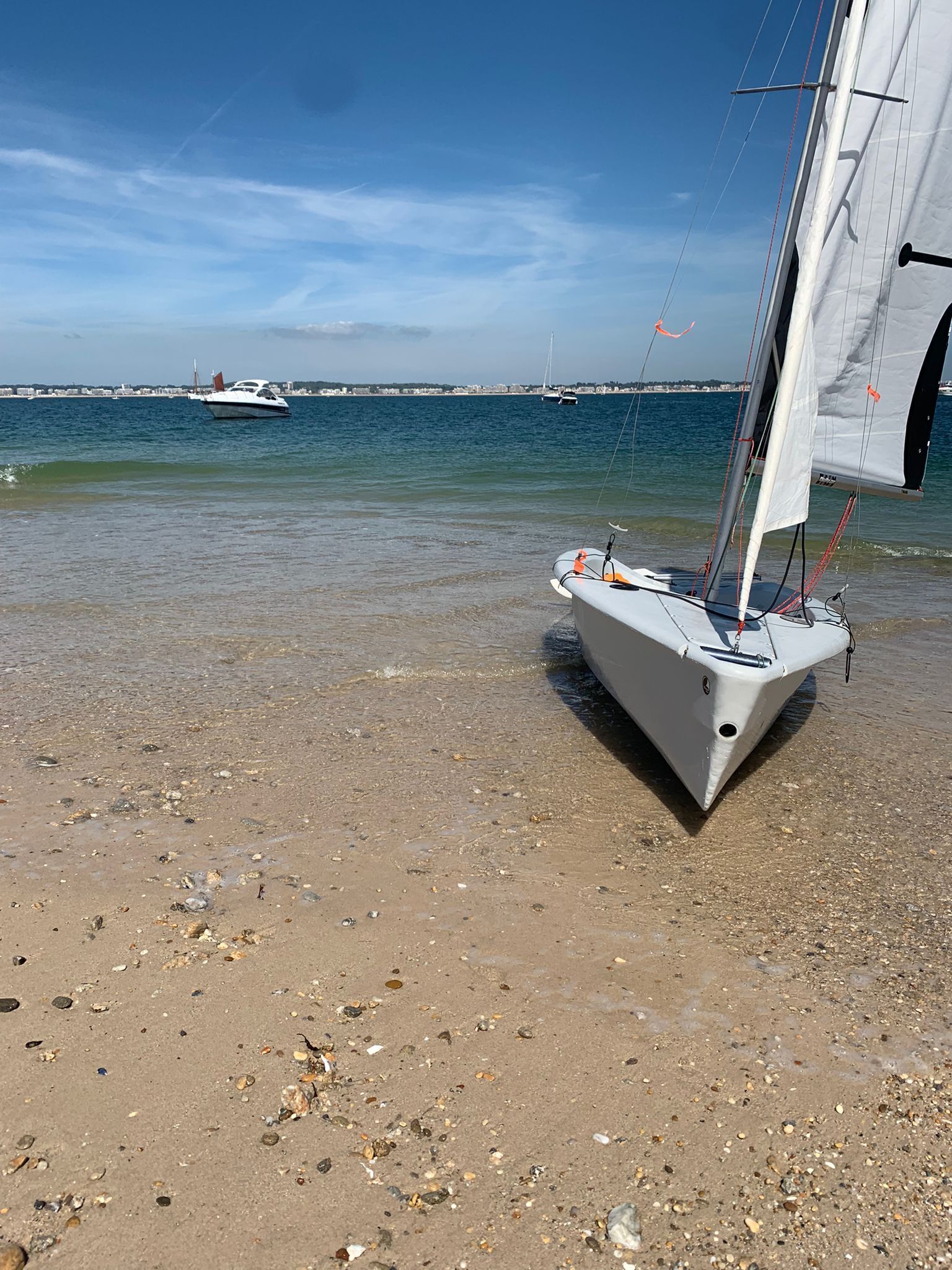 Un velero en una playa arenosa con agua tranquila y otros barcos a lo lejos.