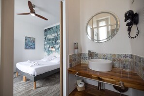 Ein Schlafzimmer mit Bett, Deckenventilator sowie einem Badezimmer mit Waschbecken, Spiegel und dekorativen Fliesen.
