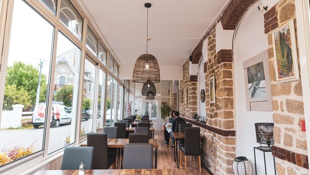 Een restaurant met een hoge plafond, grote ramen en een stenen muur. Tafels en stoelen staan klaar voor diners.