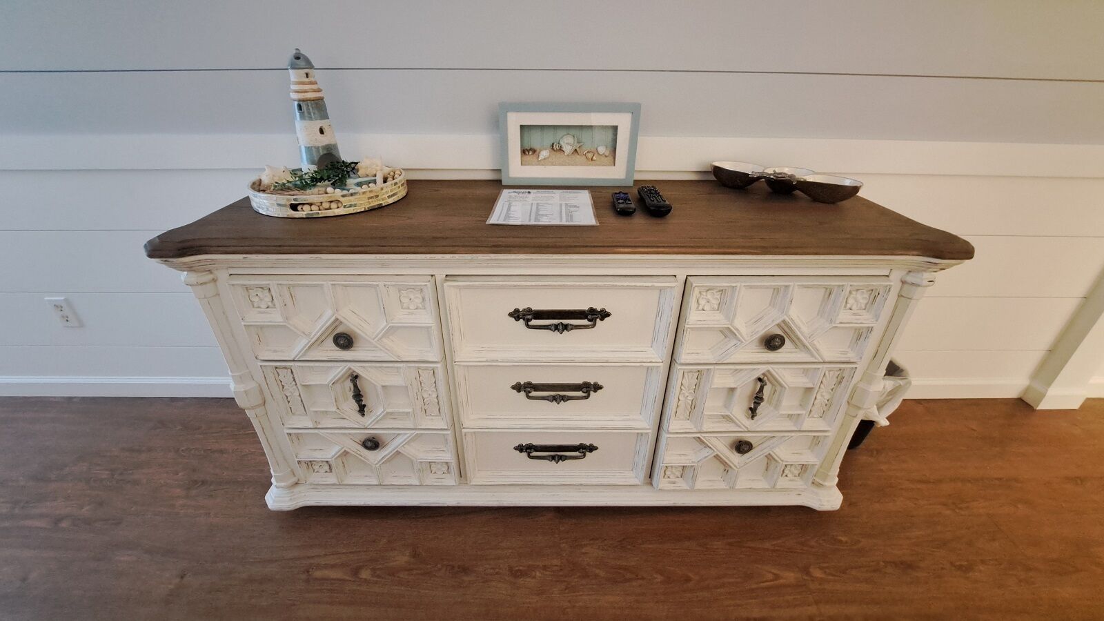 A white dresser with multiple drawers and a wooden top, featuring a lighthouse and a framed picture.