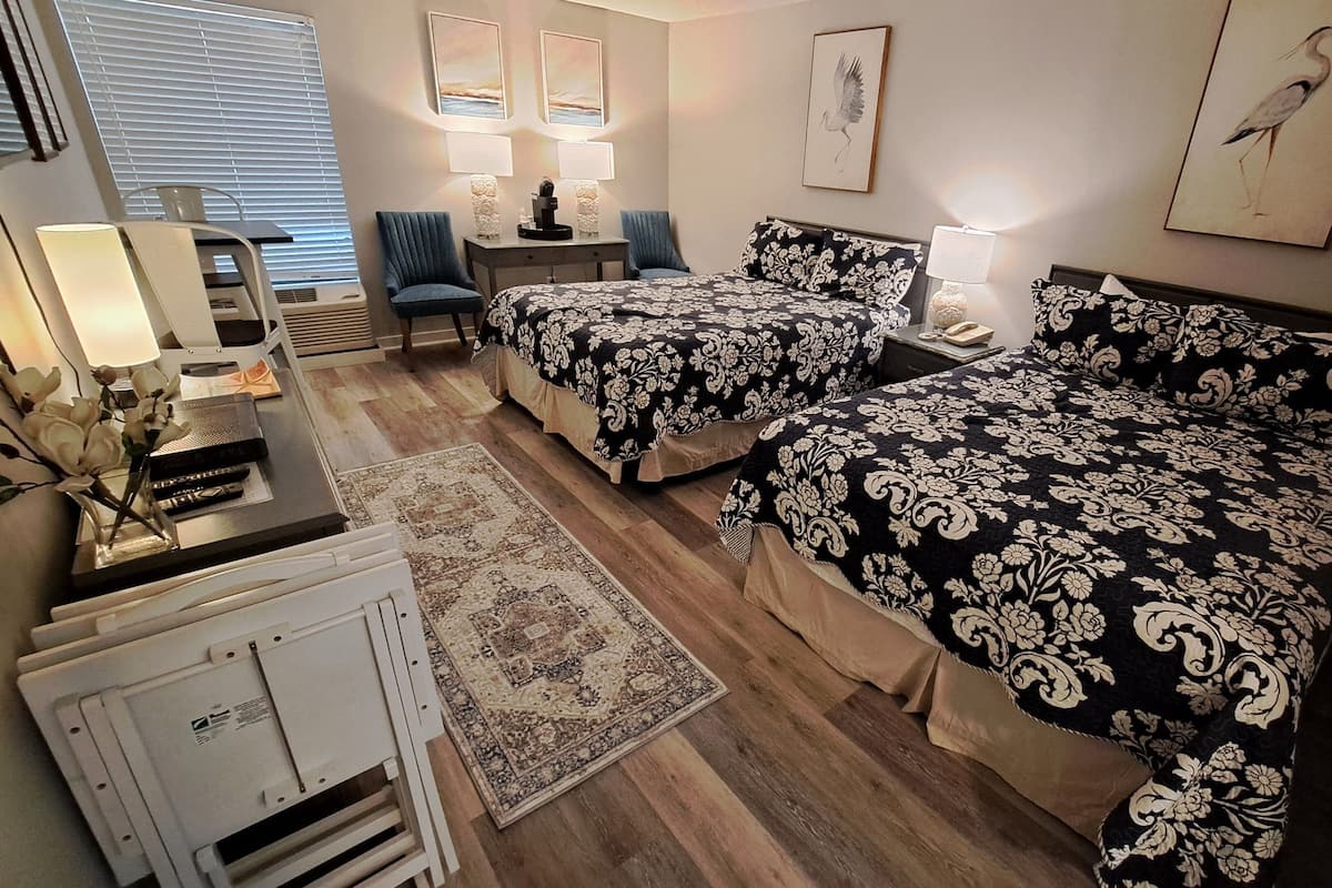 Hotellihuone, jossa on kaksi sänkyä, työpöytä, tuoli, lamppu ja taideteoksia seinillä.