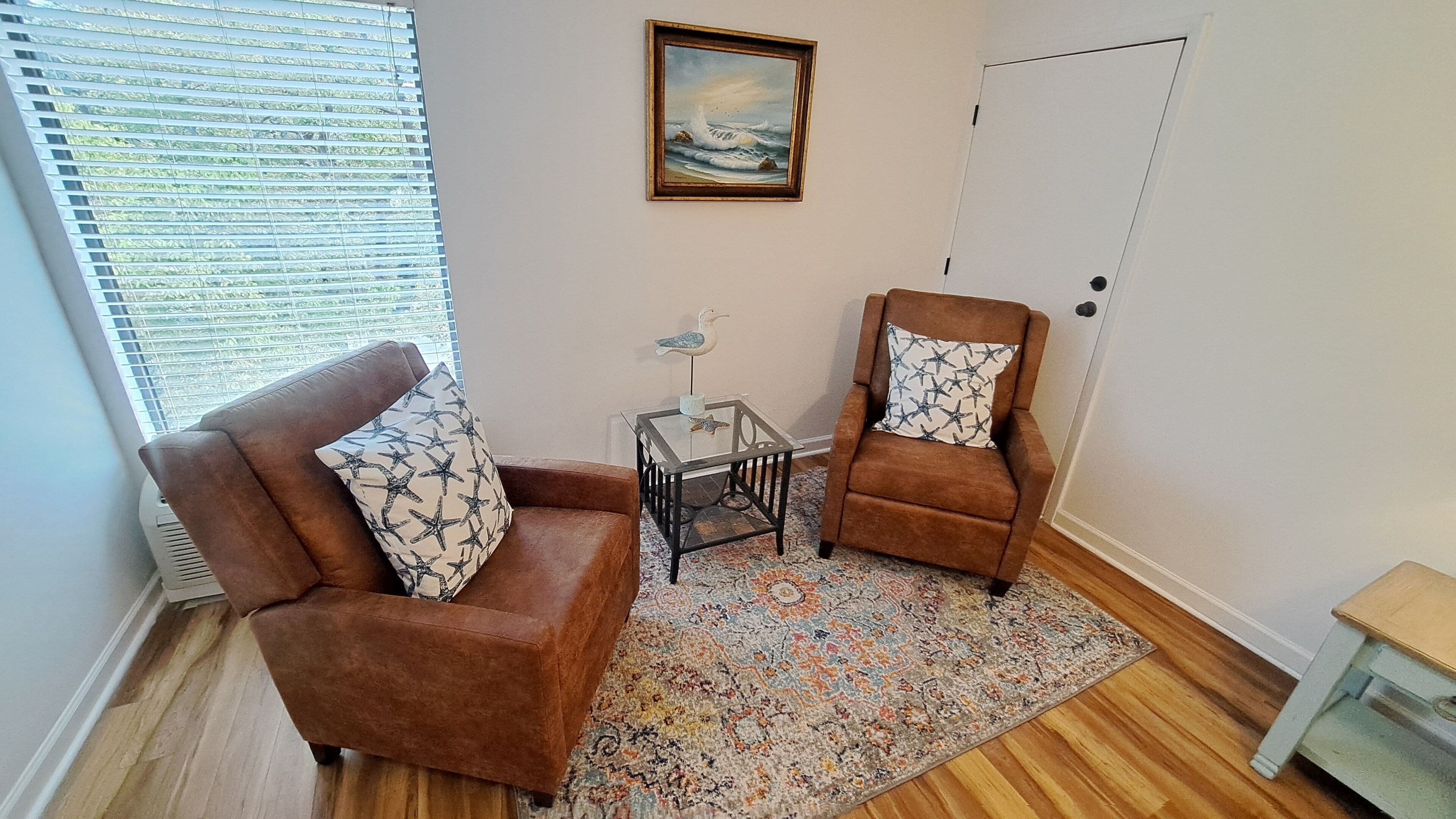 A living room with two brown armchairs, a glass coffee table, a patterned rug, a painting on the wall, and a door.